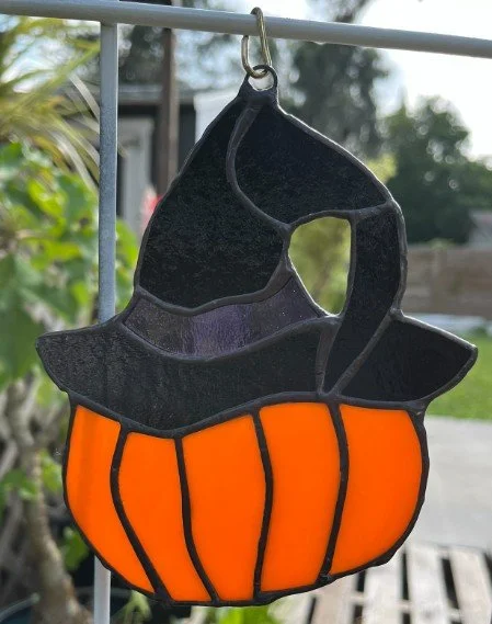 A Halloween-themed stained glass pumpkin with a black hat silhouette on top, hanging on a metal rod outdoors.