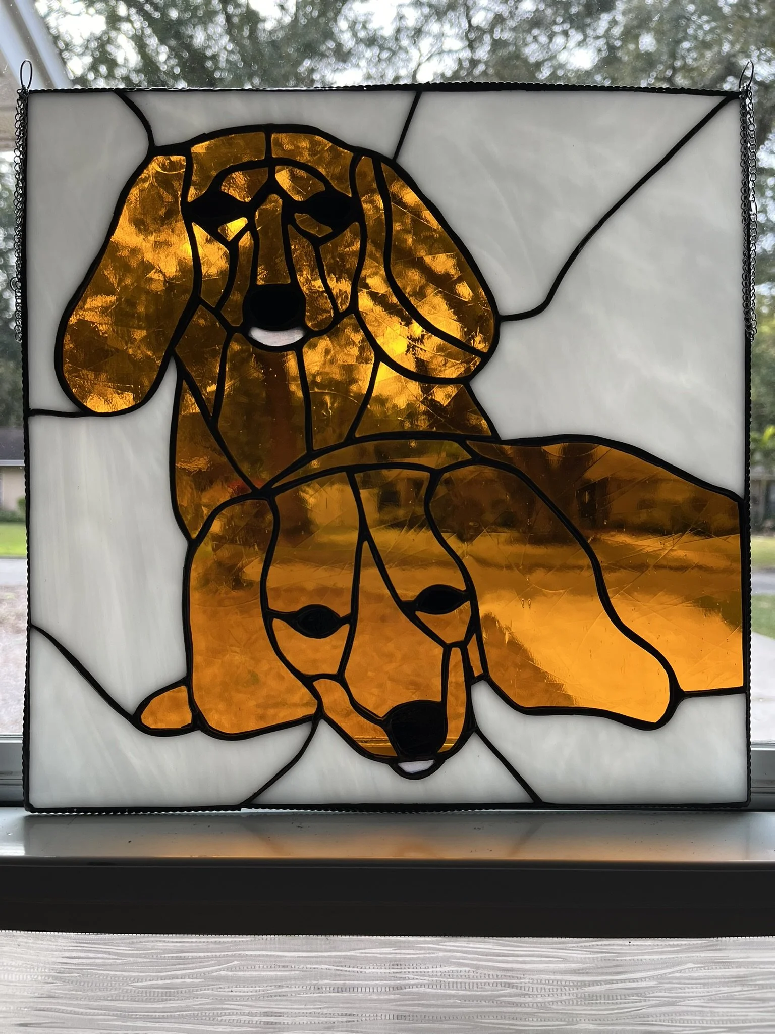 Stained glass window depicting two dachshund puppies with floppy ears, using amber and white glass with black outlines.