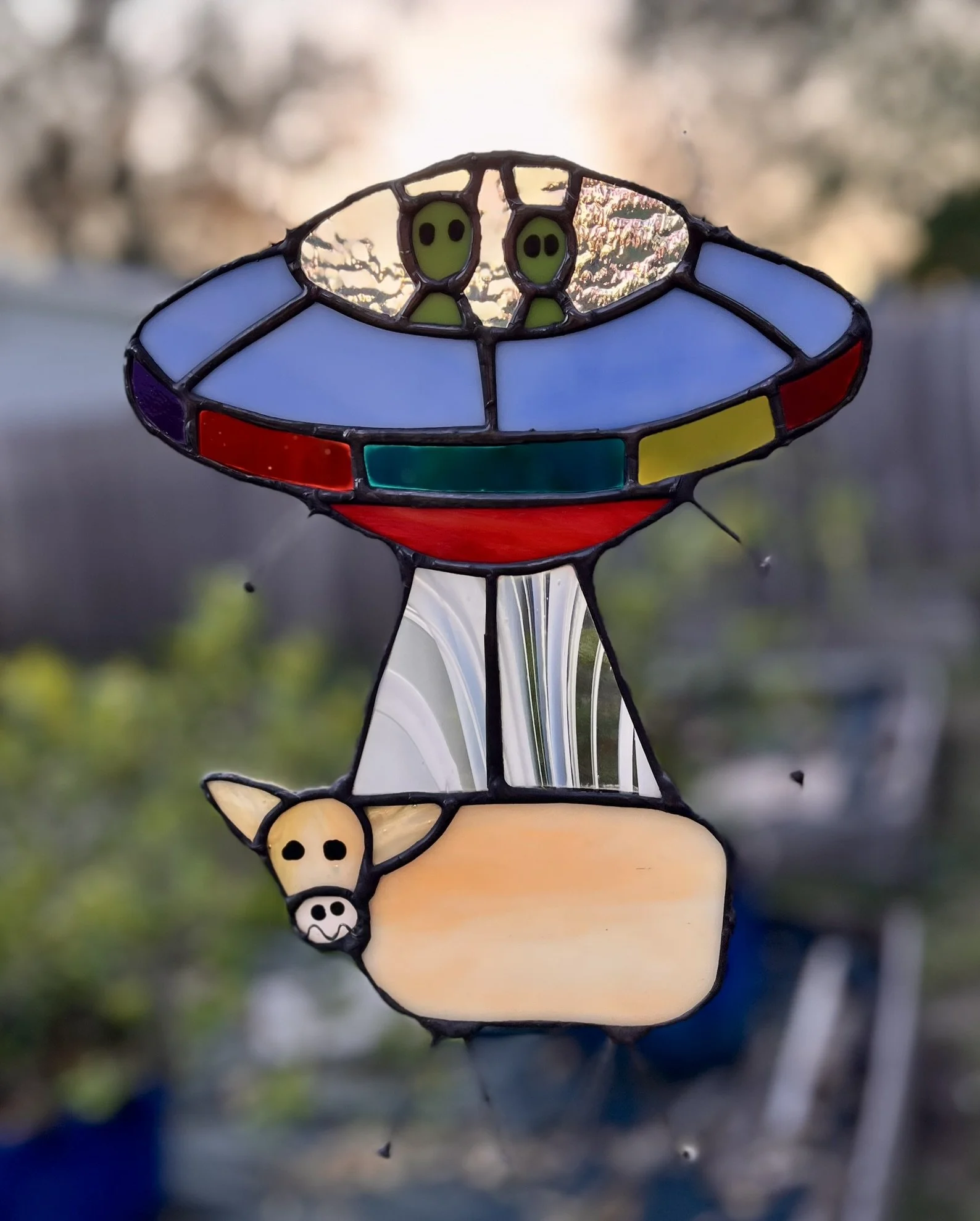 Colorful stained glass sun catcher shaped like a UFO with two green alien faces inside, and a cow with a beige face, black eyes, and white horns attached below.
