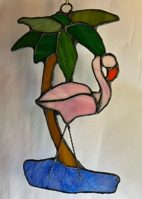A stained glass art piece depicting a pink flamingo standing on one leg on a blue pond with a palm tree with green leaves and brown trunk in the background.