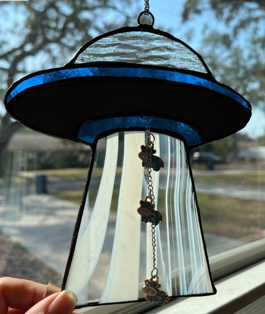 Stained glass spaceship in blue with cows with abstract swirl patterns, hanging outdoors in sunlight.