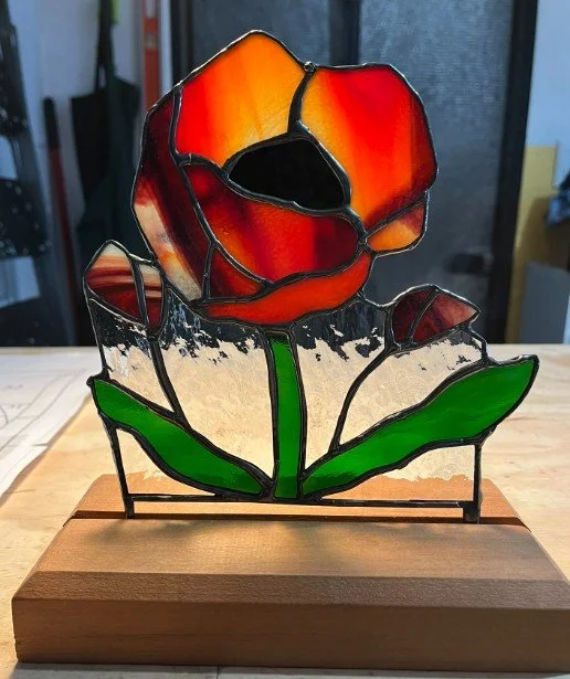 A stained glass art piece of a red and orange poppy with green leaves, mounted on a wooden base.