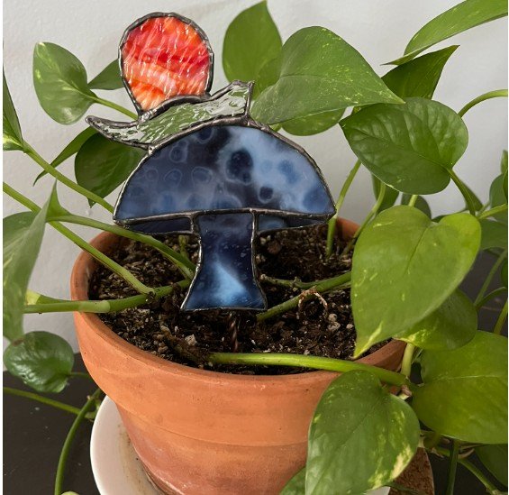 A potted pothos plant with green heart-shaped leaves. In the soil, there is a stained glass mushroom decoration with a red strawberry on top.