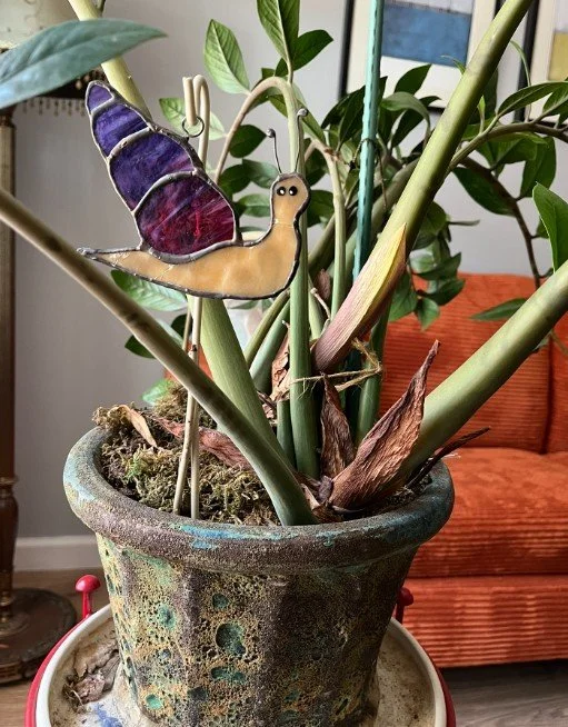 Decorative snail ornaments in a plant pot with green leafy plants and a background of a sofa.