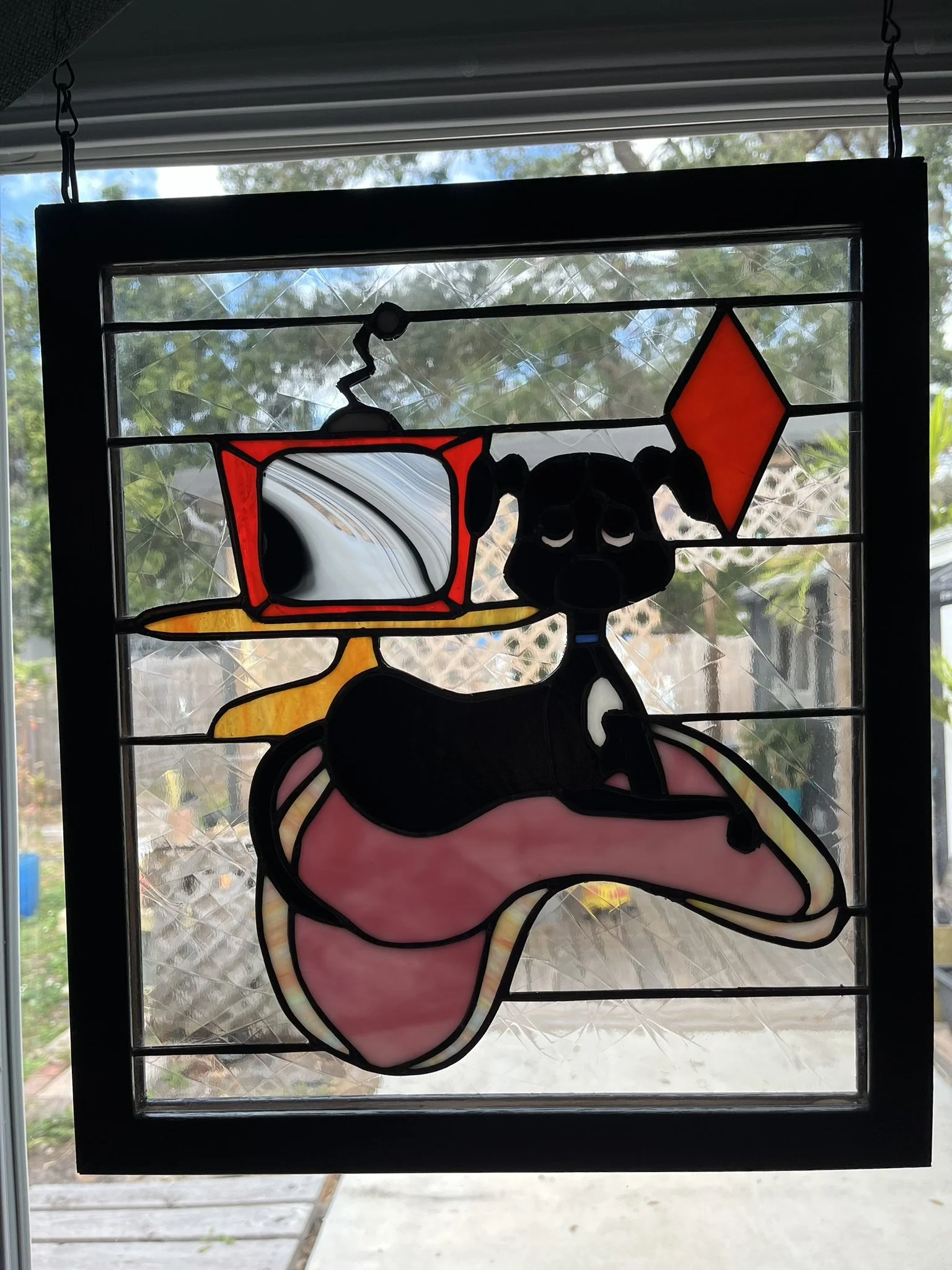 Stained glass window featuring a black puppy lying on a pink rug with an orange diamond-shape in the background.