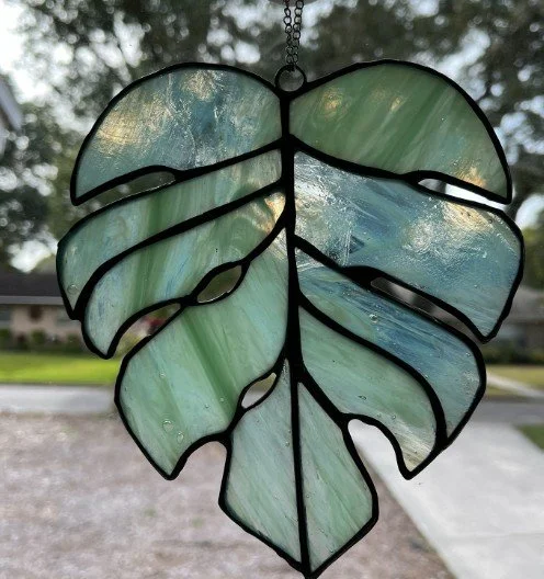 Stained glass sun catcher in the shape of a Monstera leaf with greenish-blue hues, hanging outdoors against trees and a cloudy sky.