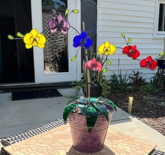 Colorful stained glass Birthstone Orchid in a purple flower pot on a table outside a house.