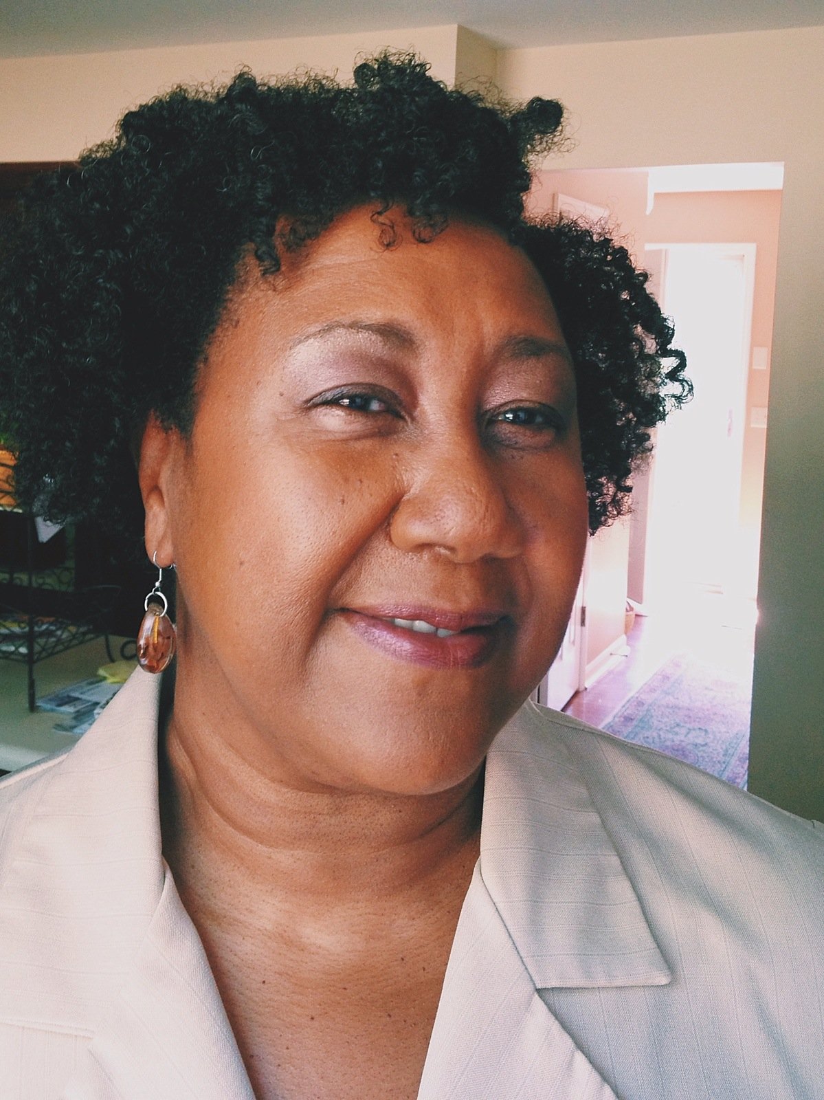 Close-up of a woman with curly black hair, wearing earrings and a light-colored blazer, smiling inside a well-lit home.
