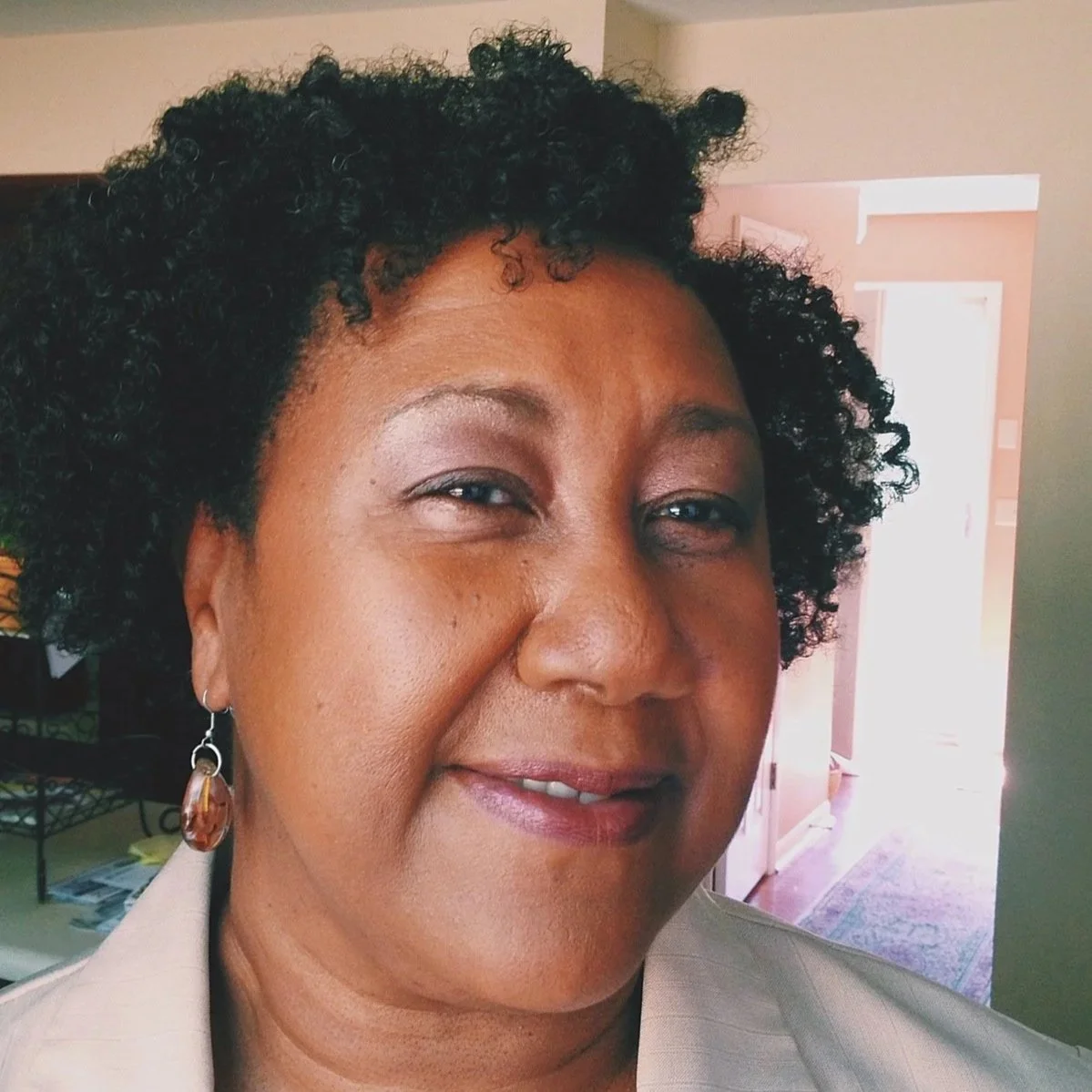 Close-up of a smiling woman with curly black hair, wearing a beige blazer and a large amber earring, standing inside a home with a doorway and hallway visible in the background.