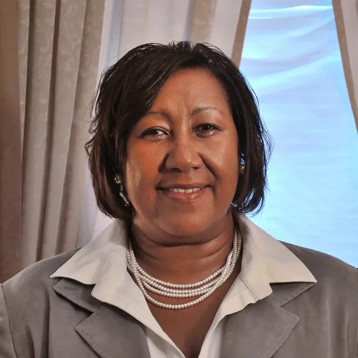 A middle-aged woman with dark hair styled in a bob, wearing pearl earrings, layered pearl necklaces, a white shirt, and a light gray blazer, standing indoors with beige curtains and a window behind her.