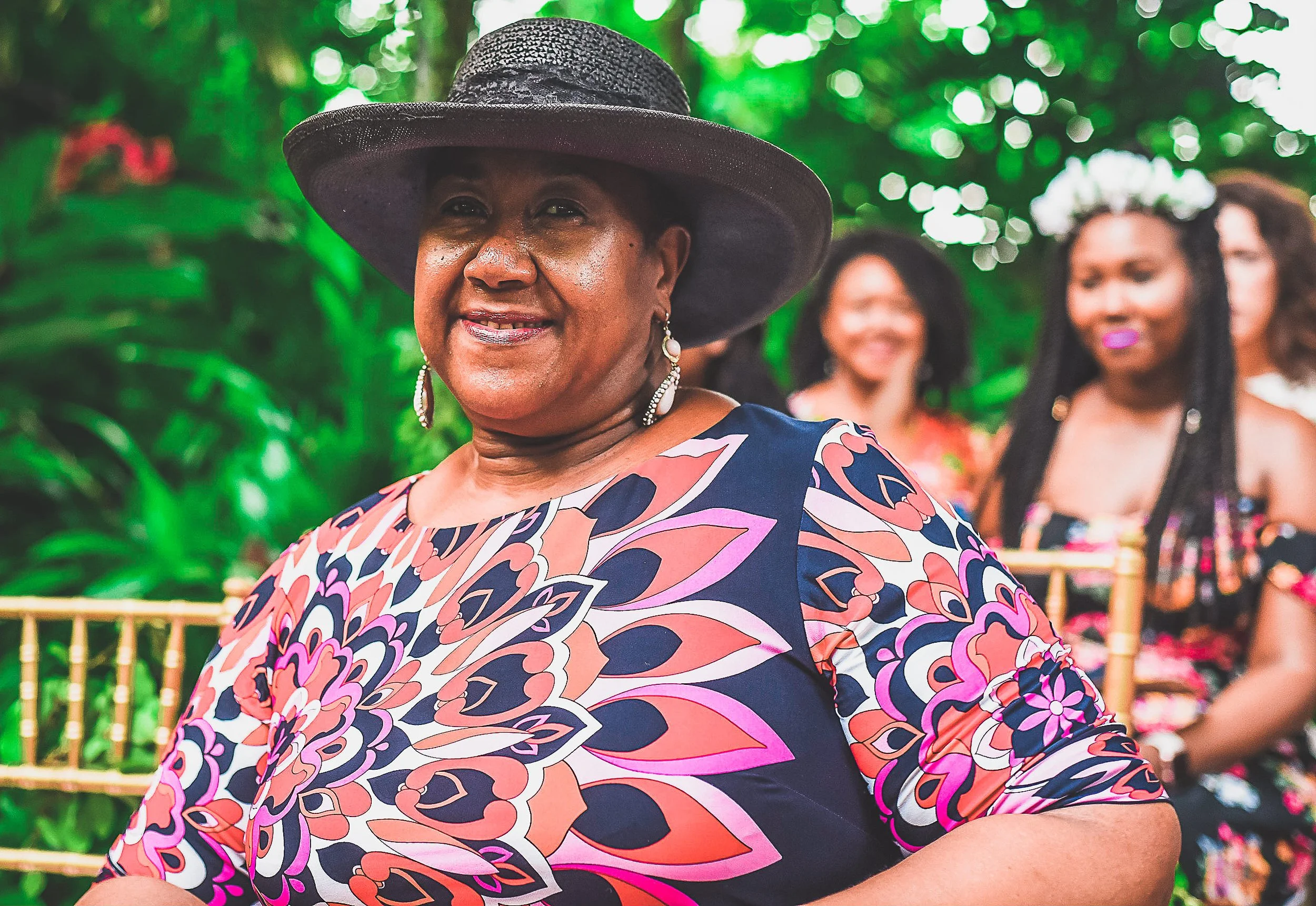 A smiling woman wearing a wide-brimmed hat and a colorful floral dress, standing outdoors with green foliage in the background, with several women sitting behind her.