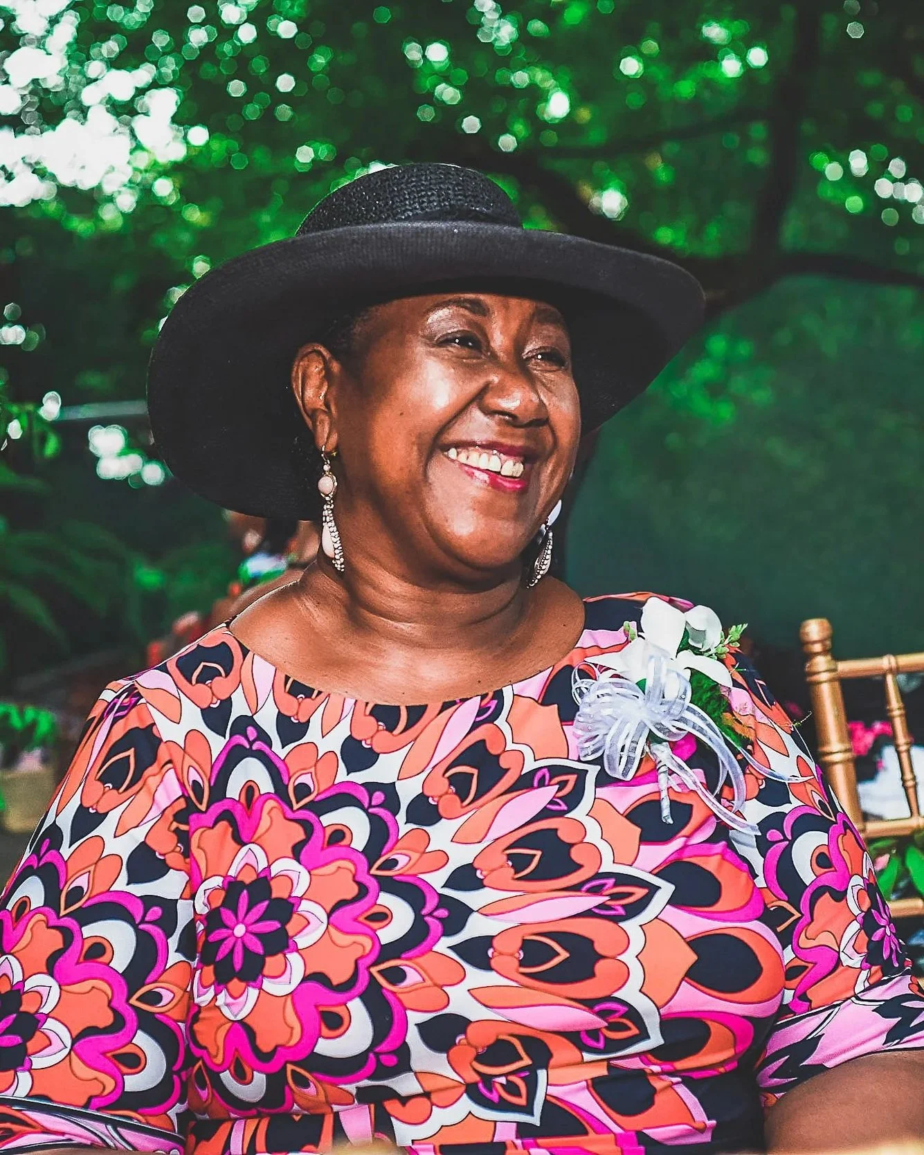 Smiling woman wearing a large black hat, floral dress, and earrings, sitting outdoors with a blurred green background.