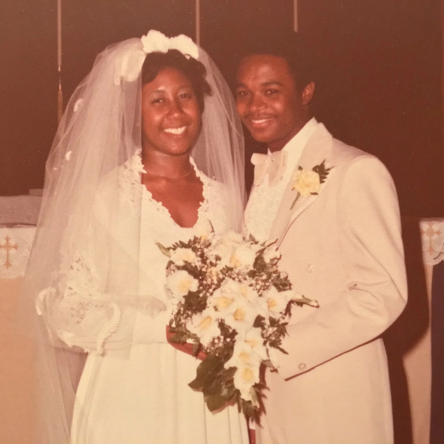 A bride and groom in wedding attire, smiling and standing close together indoors, with the bride holding a bouquet of white flowers.