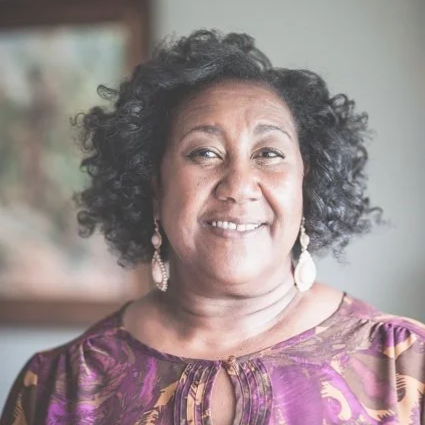 A smiling woman with curly black hair wearing a purple patterned top and earrings.