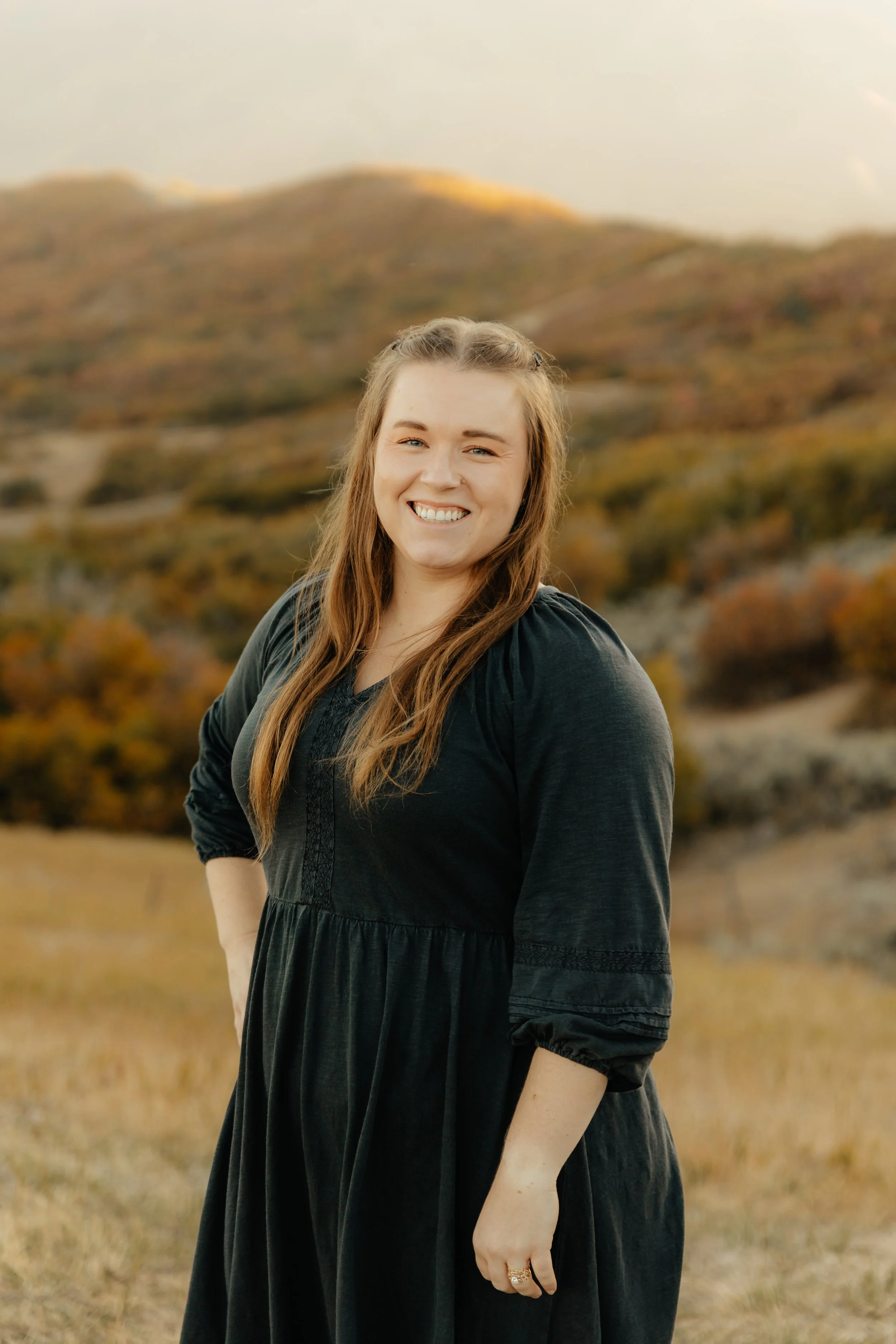 A young woman with long brown hair smiling in an outdoor landscape with mountains and autumn foliage.