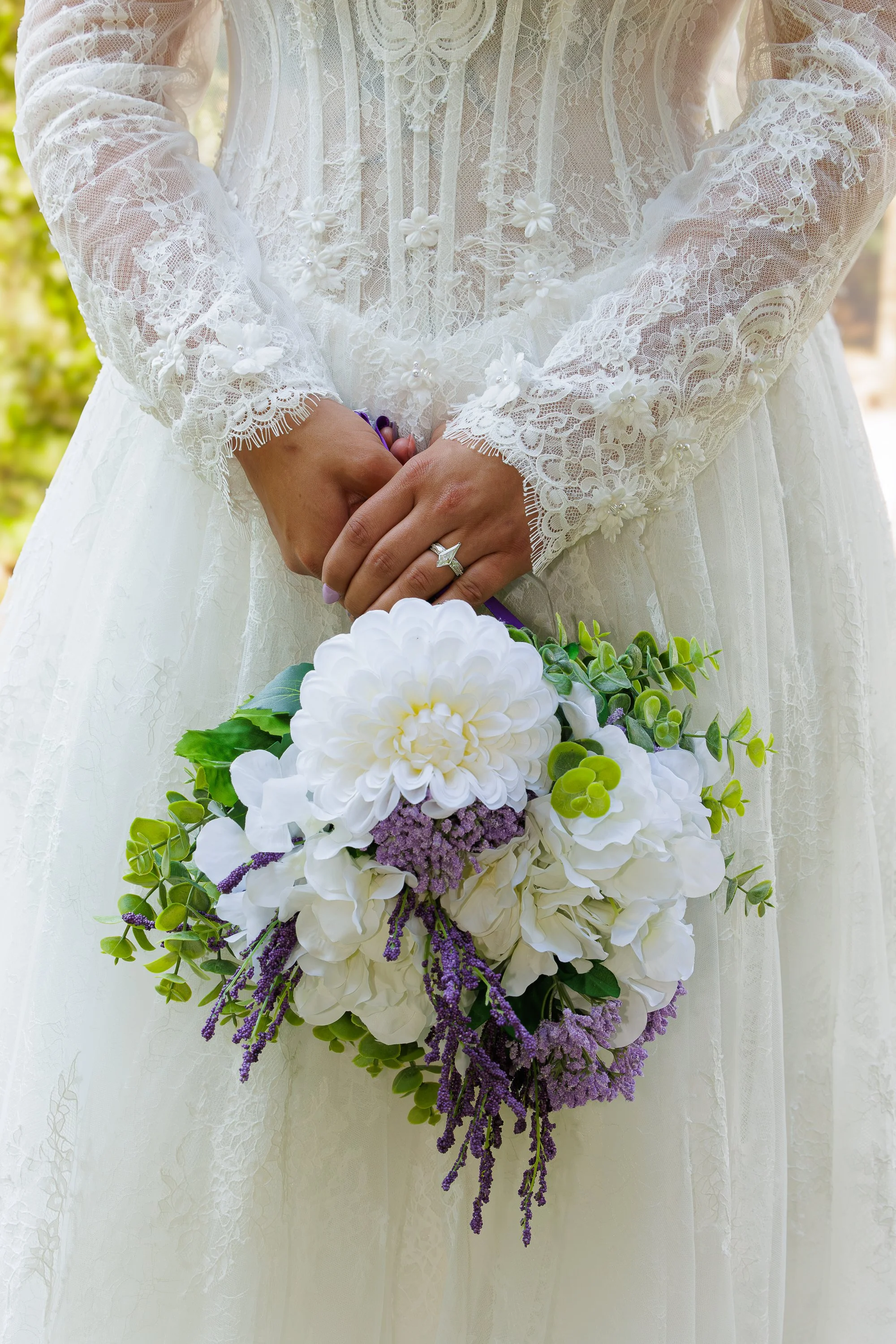 A person in a wedding dress holding a bouquet of white and purple flowers, with lace details on the dress and long sleeves.