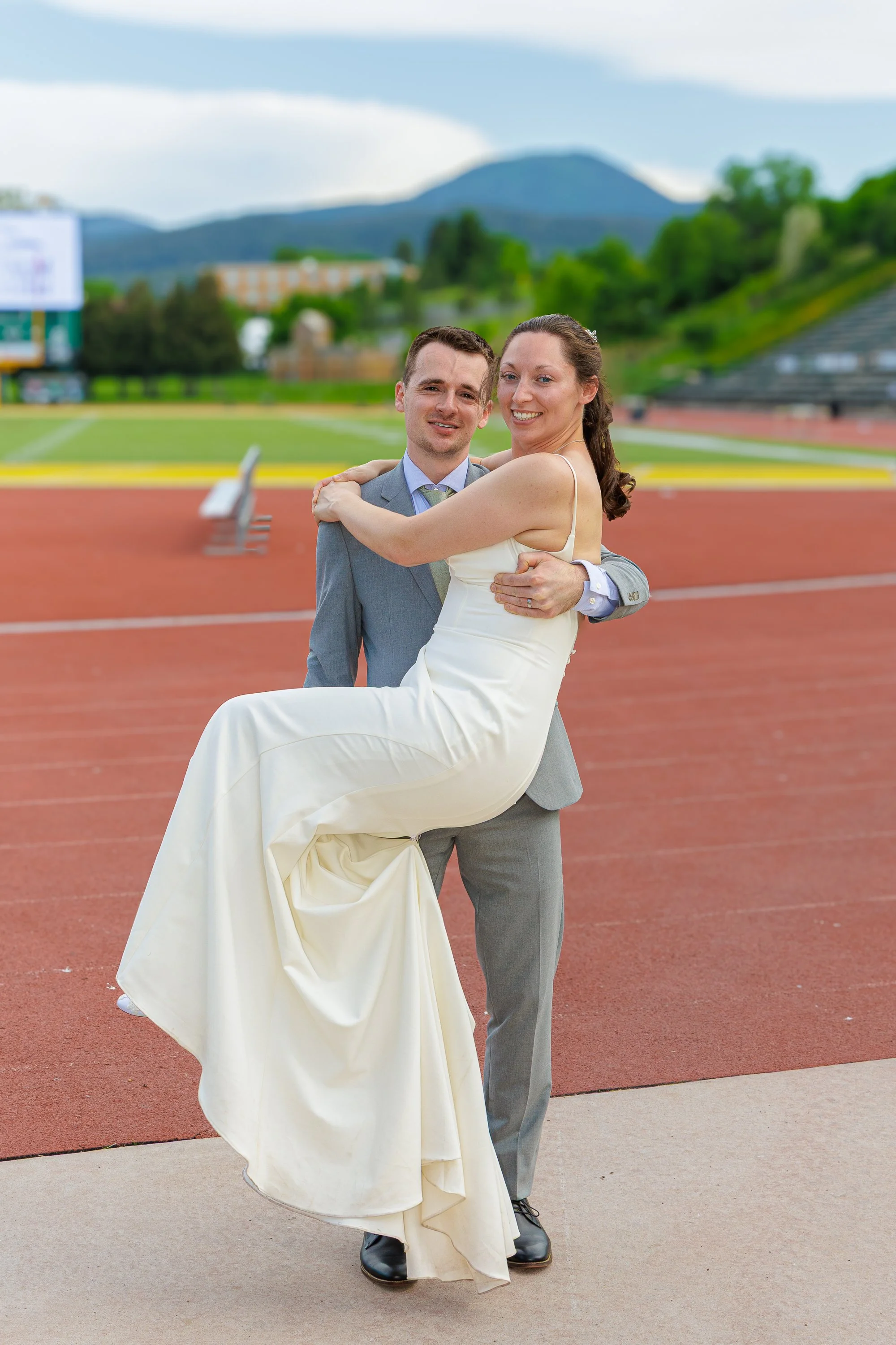 A bride and groom at an outdoor track field, with the groom holding the bride in his arms. The bride is wearing a white wedding dress, and the groom is dressed in a gray suit. Mountains and a blue sky with some clouds are visible in the background.