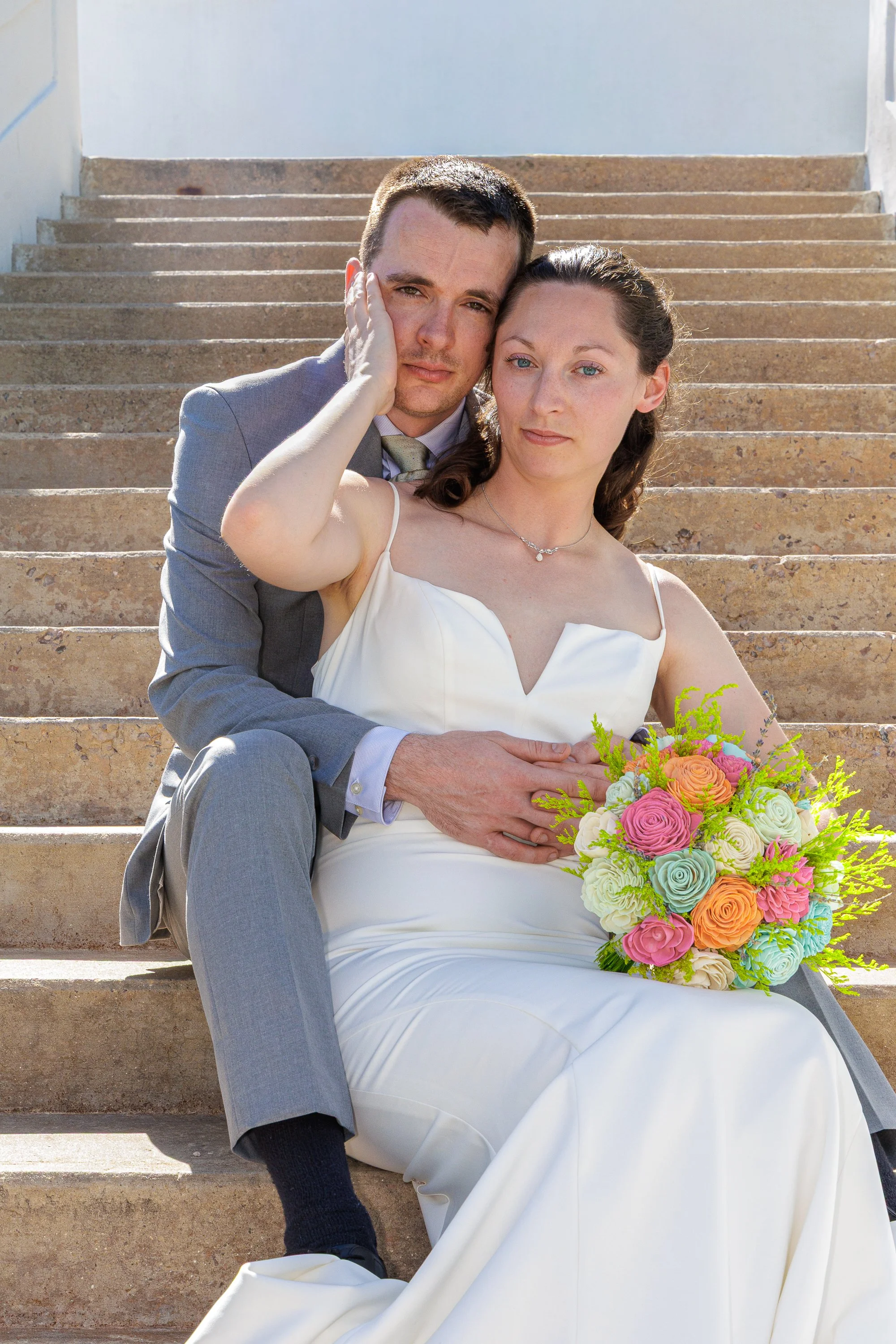 A newlywed couple sitting on outdoor steps, with the groom in a gray suit and the bride in a white wedding dress holding a colorful bouquet of flowers.