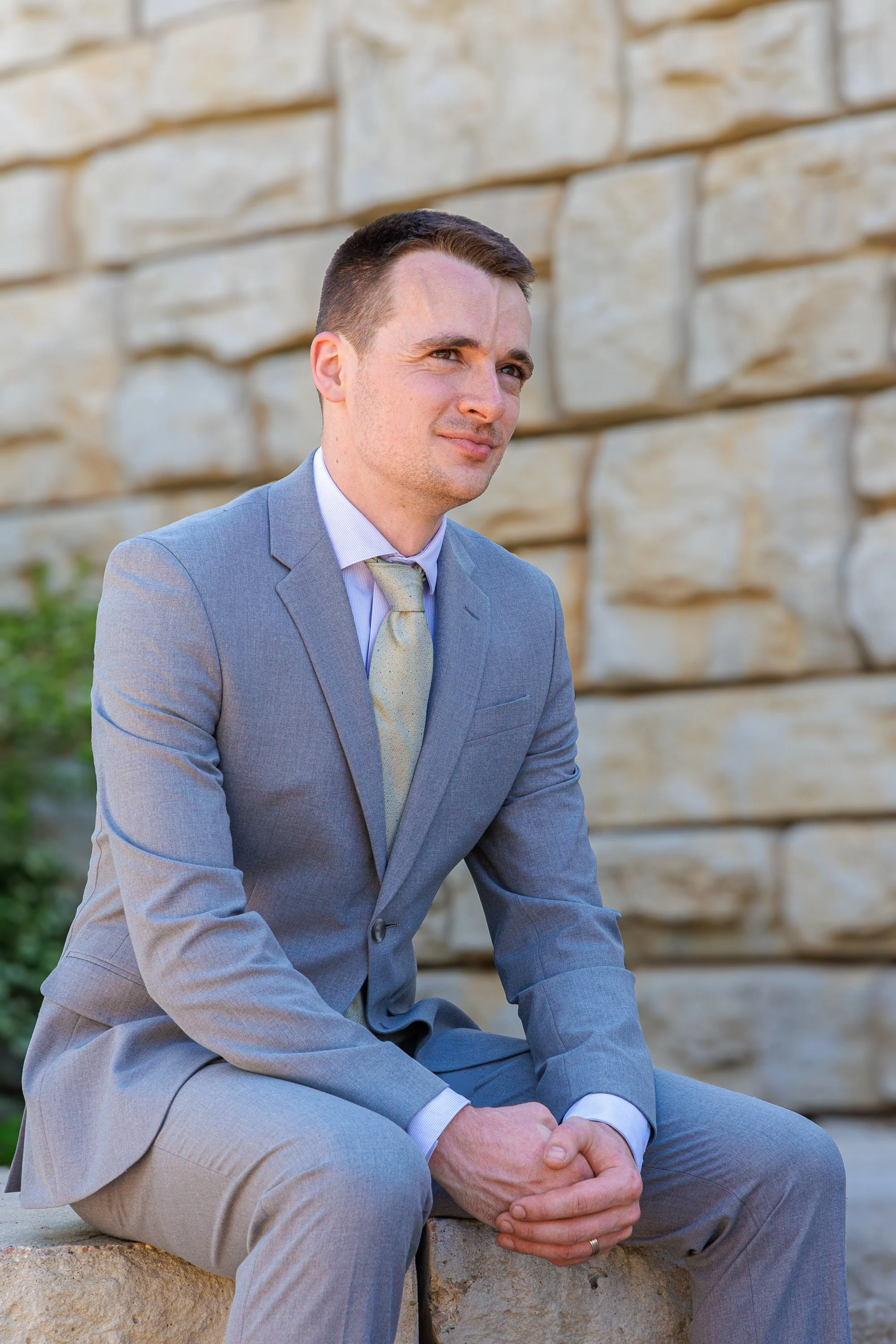 A man in a light gray suit and tie sitting on a stone ledge in front of a beige brick wall with greenery nearby, looking to the side with a thoughtful expression.