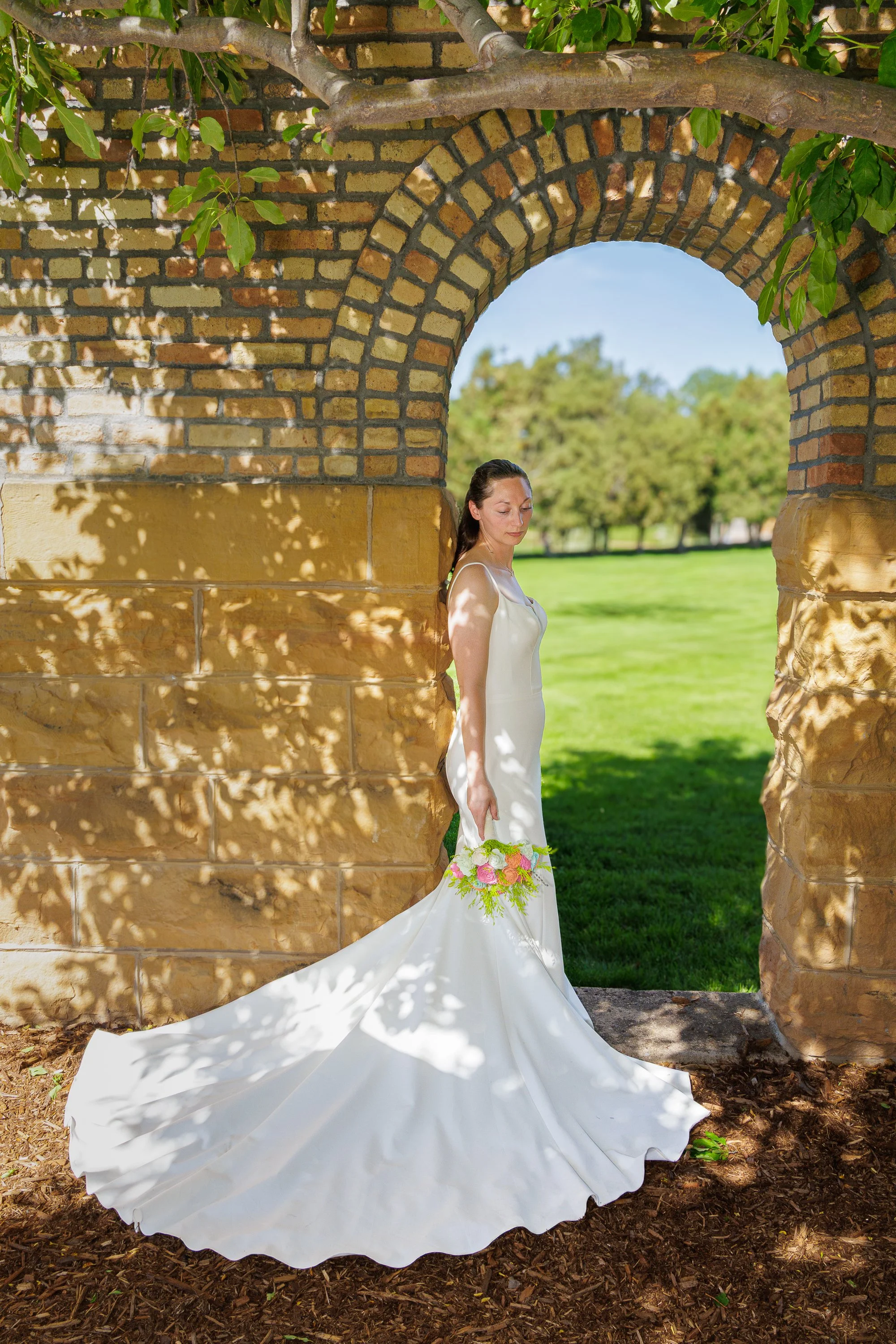 A woman in a white wedding dress holding a bouquet of flowers standing under a brick archway in a park with green grass and trees.