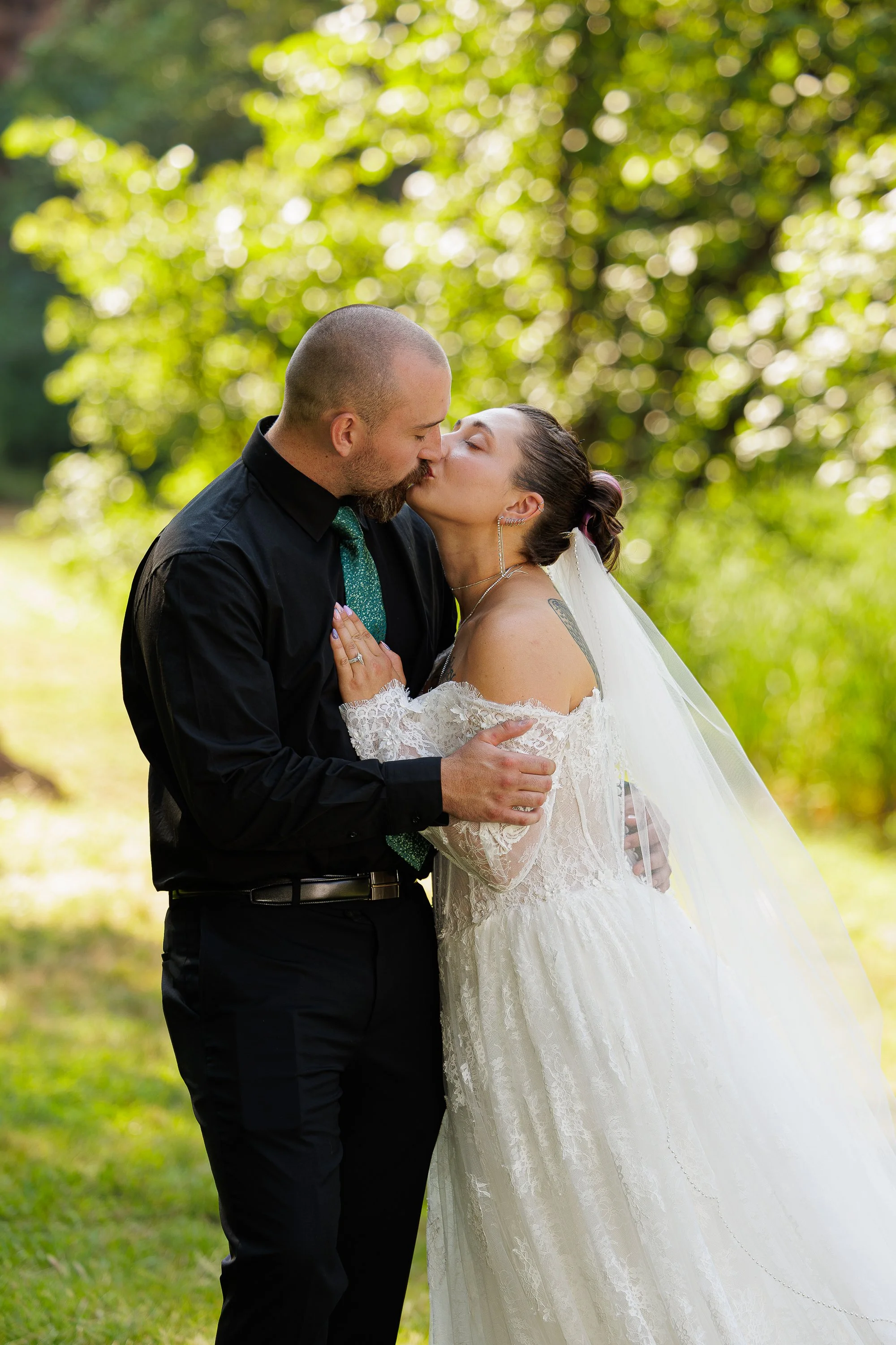 A couple kissing outdoors during their wedding, with the bride in a white lace dress and veil, and the groom in a black shirt and tie, surrounded by green trees with sunlight filtering through.