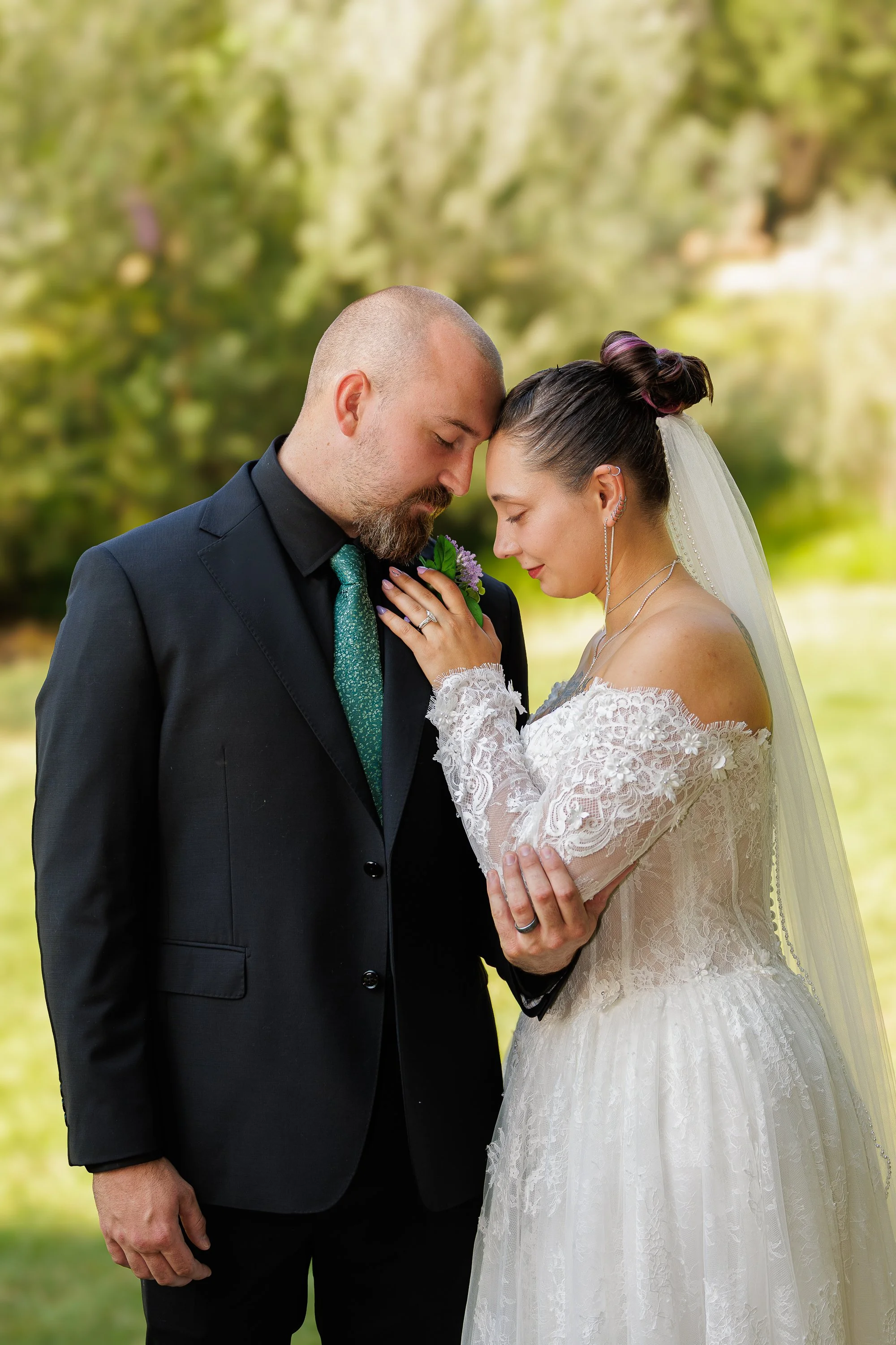 A couple dressed in wedding attire, with eyes closed and foreheads touching, standing outdoors in a natural setting with blurred green foliage in the background.