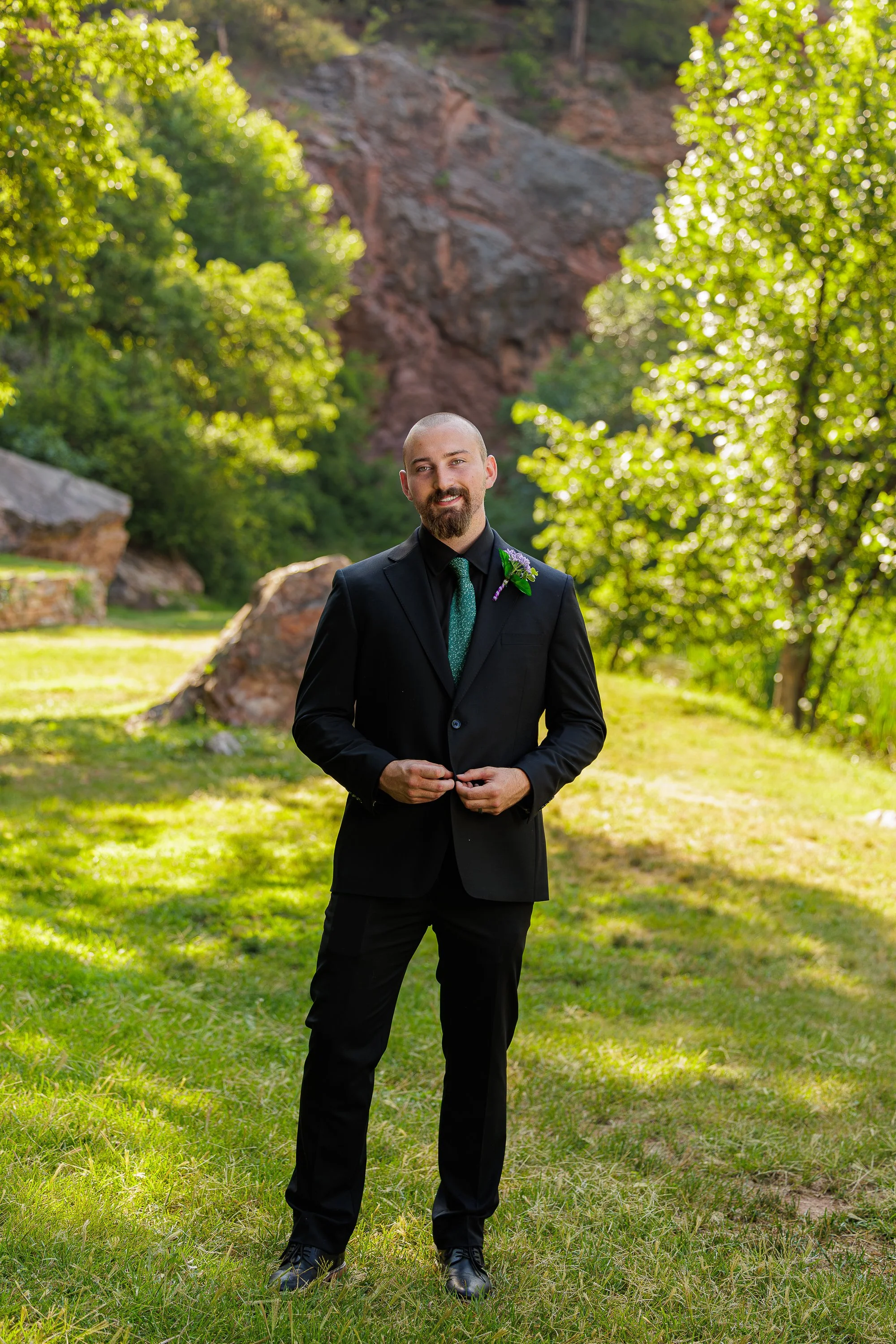 A man dressed in a black suit with a green tie and boutonniere standing outdoors on green grass, smiling, with greenery and rocks in the background.