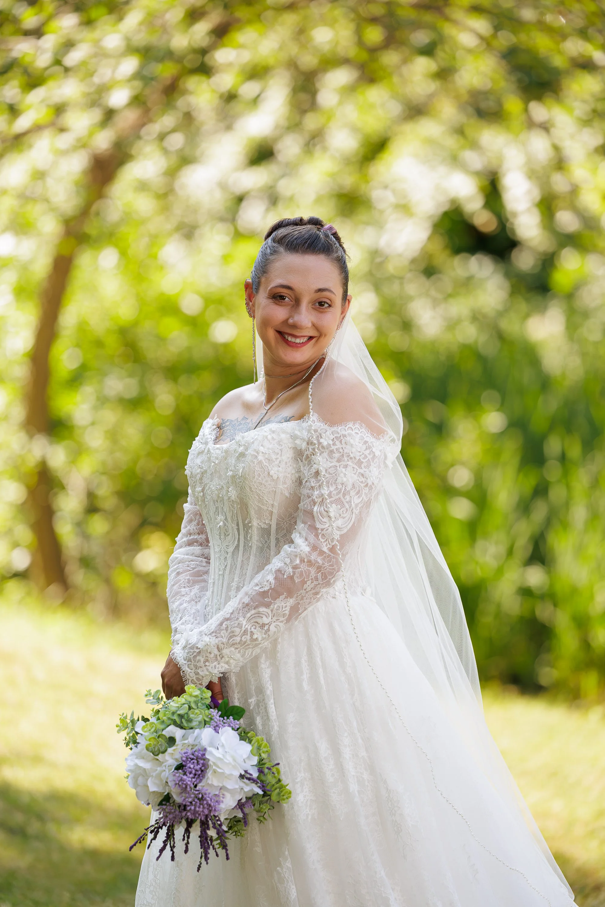 A woman in a wedding dress holding a bouquet of purple, white, and green flowers in the Black Hills of South Dakota, with leafy trees in the background.