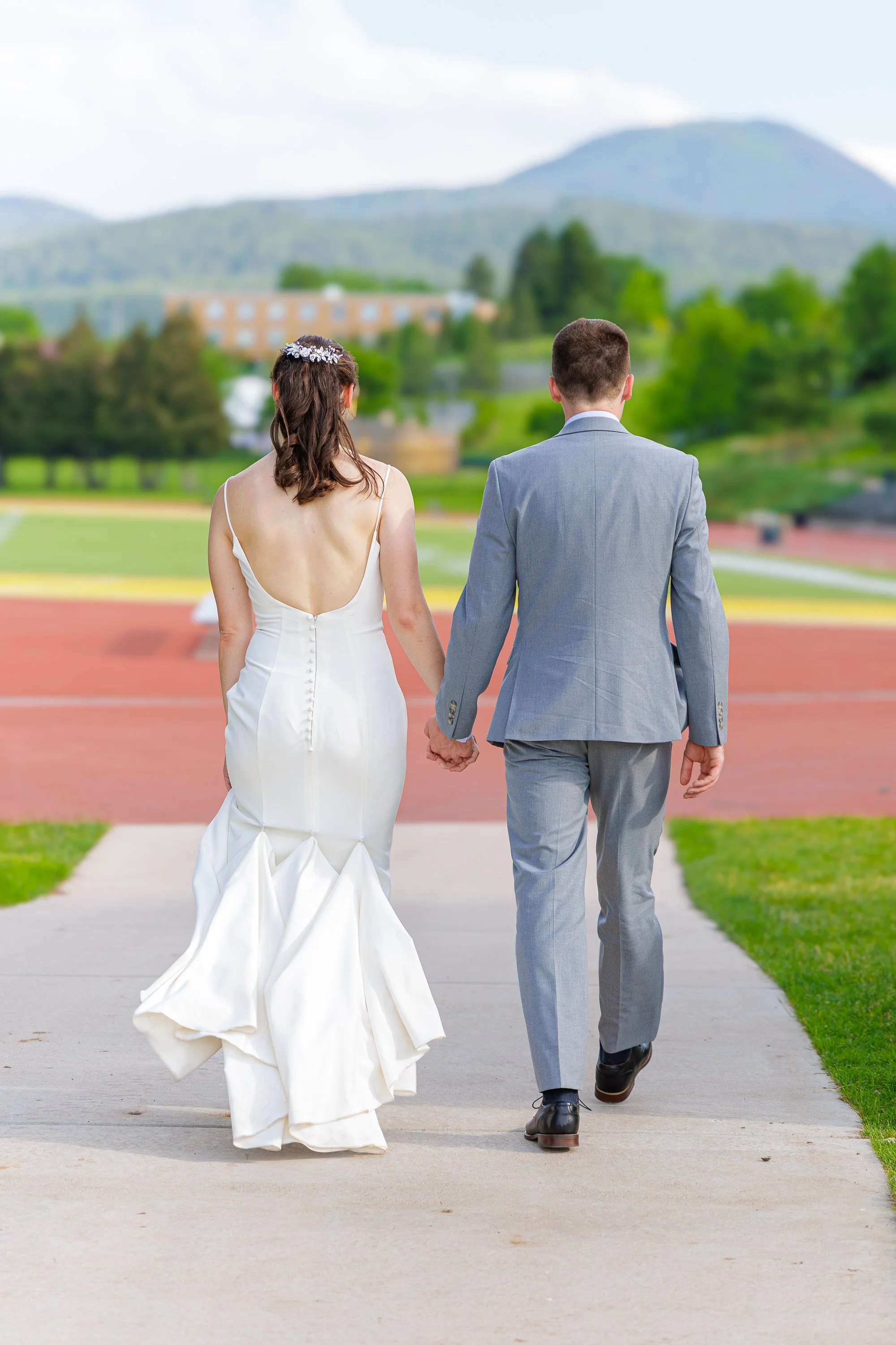 A bride and groom walking hand in hand outdoors, seen from behind, with lush green trees and mountains in the background.