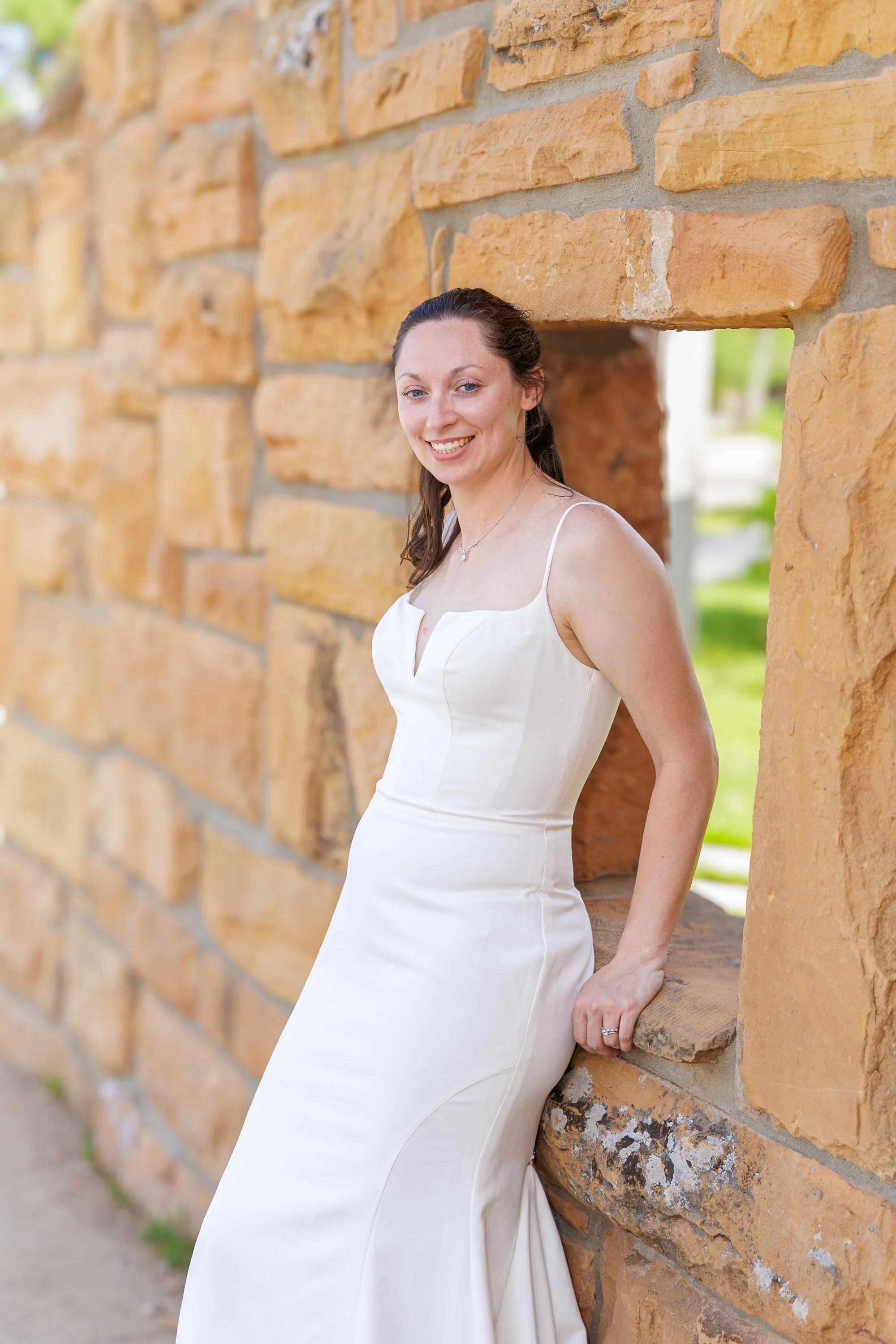 A woman in a white dress smiling, leaning against a stone wall with greenery in the background.