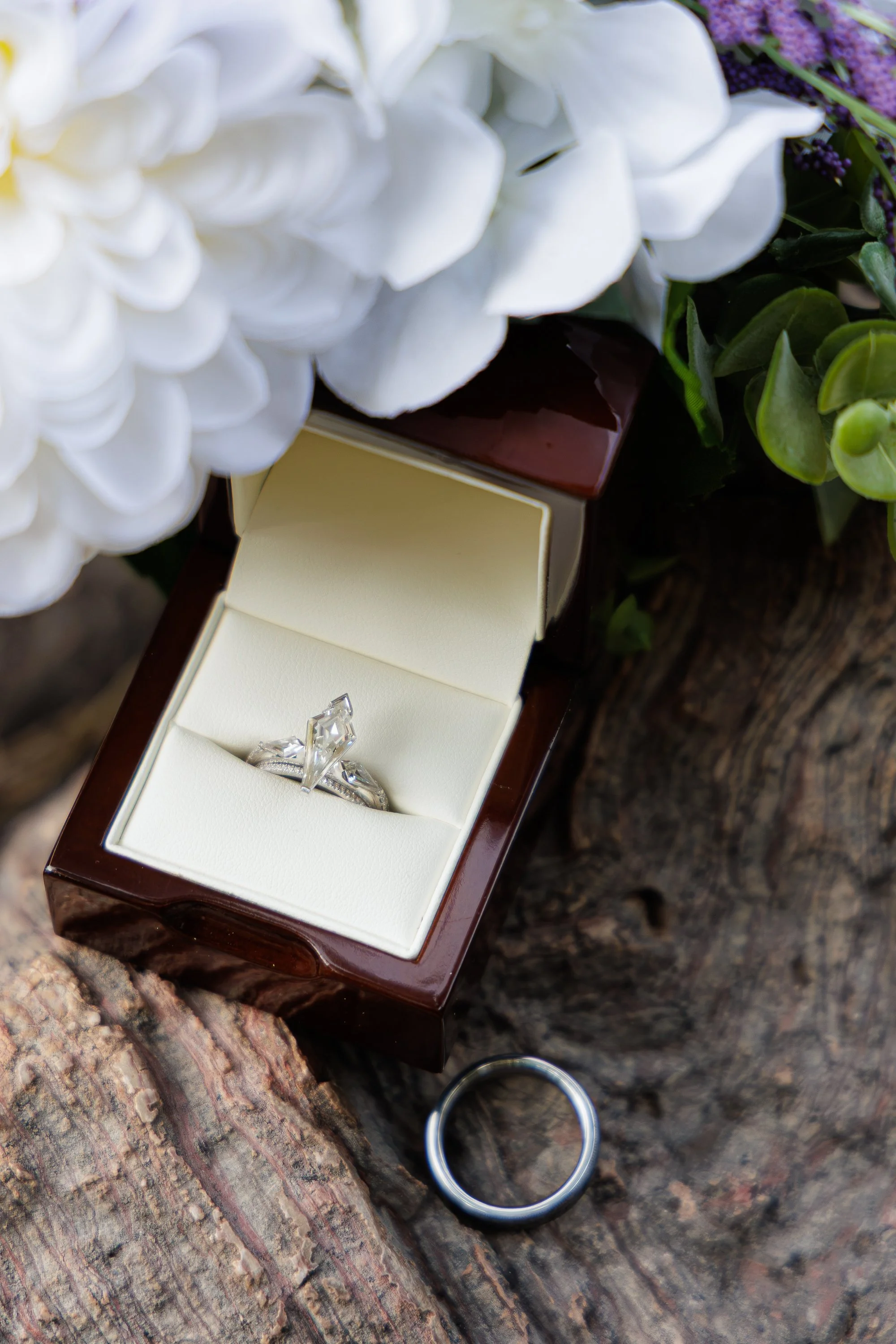 Close-up of a jewelry box with a diamond ring inside, surrounded by white and purple flowers and greenery on a wooden surface.
