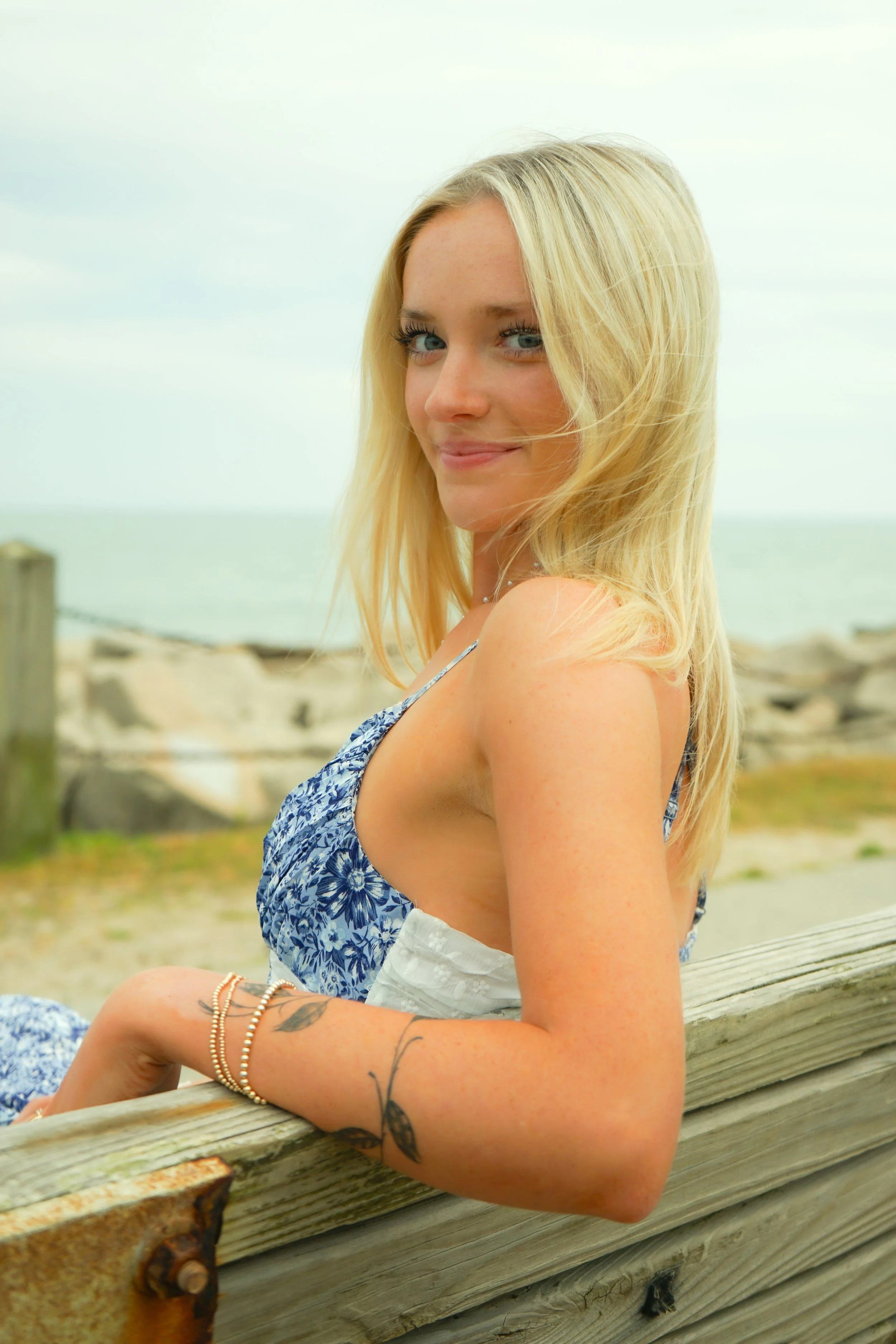 A blonde woman with blue eyes, smiling, sitting outdoors near a beach, wearing a blue floral sleeveless top, with a tattoo on her forearm and bracelets.