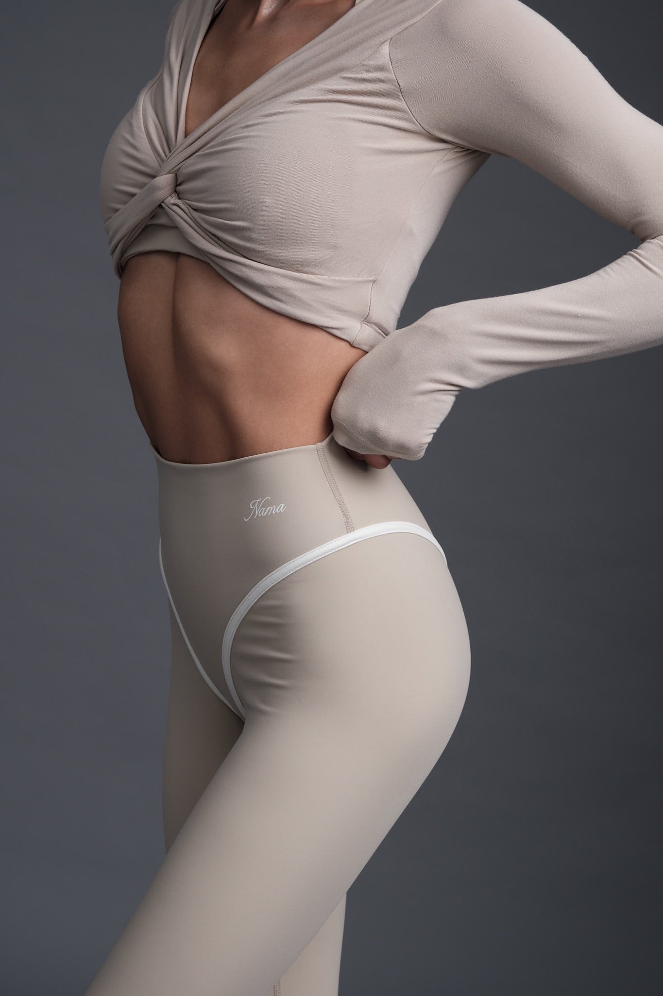 A woman from the neck down wearing a beige long-sleeve top with a twisted knot at the chest and beige high-waisted leggings with the word 'Nana' on the waistband, standing against a dark gray background.