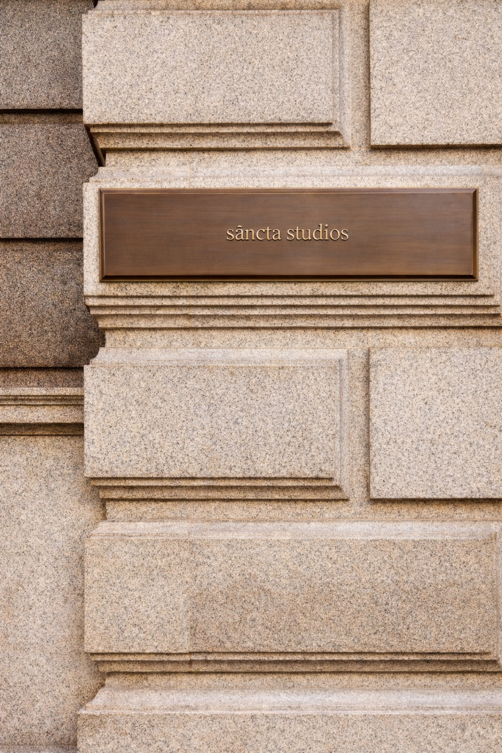 A brick wall with a bronze plaque that reads 'sáncta studios' mounted on it.