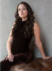 Woman with long dark hair sitting on a leather couch, wearing a black sleeveless top, against a gray wall.