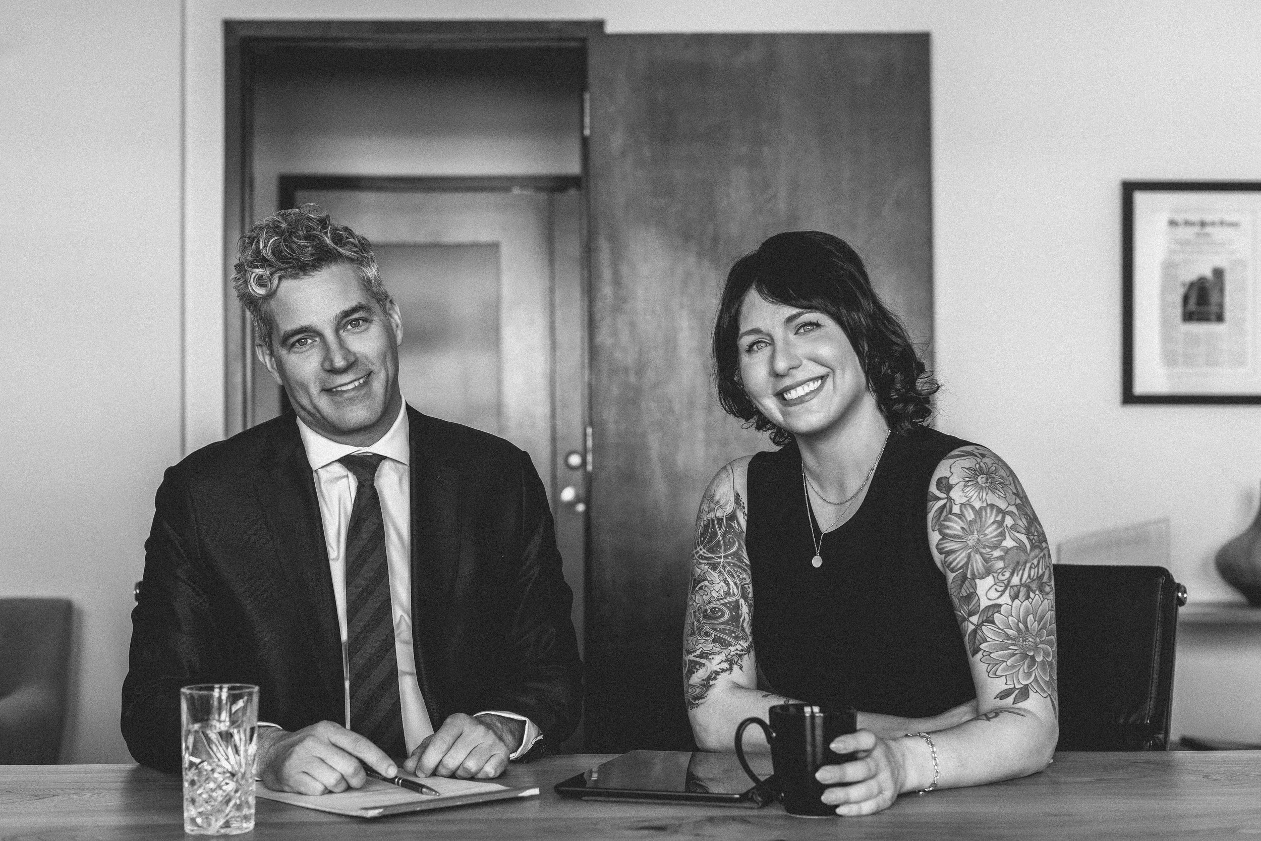 Black and white photo of Partners Dan Curry and Sarah Duggan sitting at a table in an office, smiling at the camera.