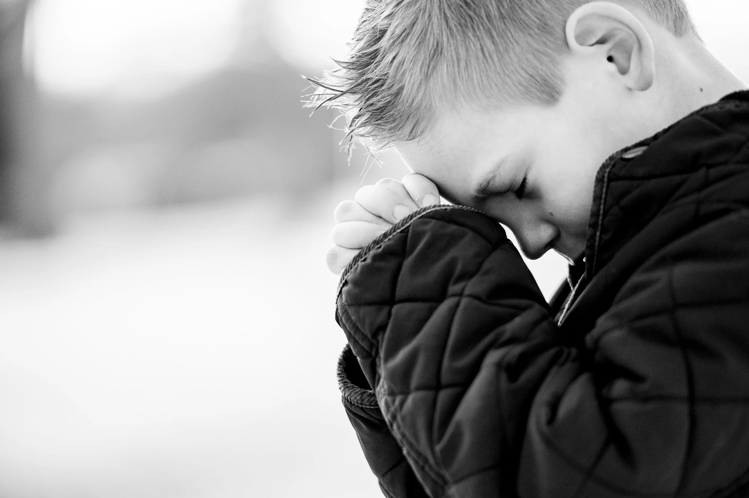 A young boy with short light hair, wearing a quilted jacket, has his hands clasped together and eyes closed, appearing to be visibility distraught.