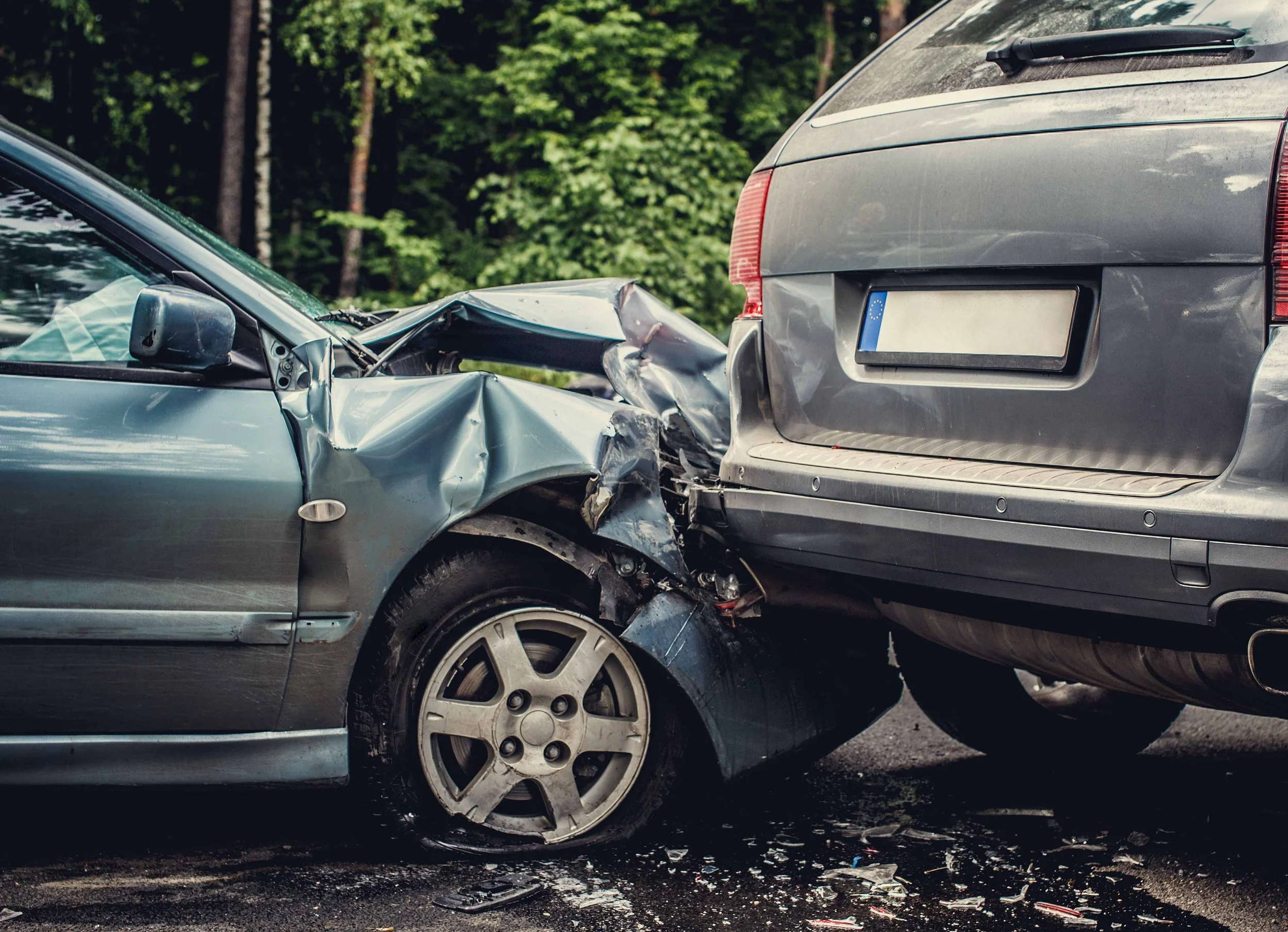 A car crash showing a silver vehicle with front-end damage crashing into the rear of a gray car on a road, with debris scattered on the ground and trees in the background.