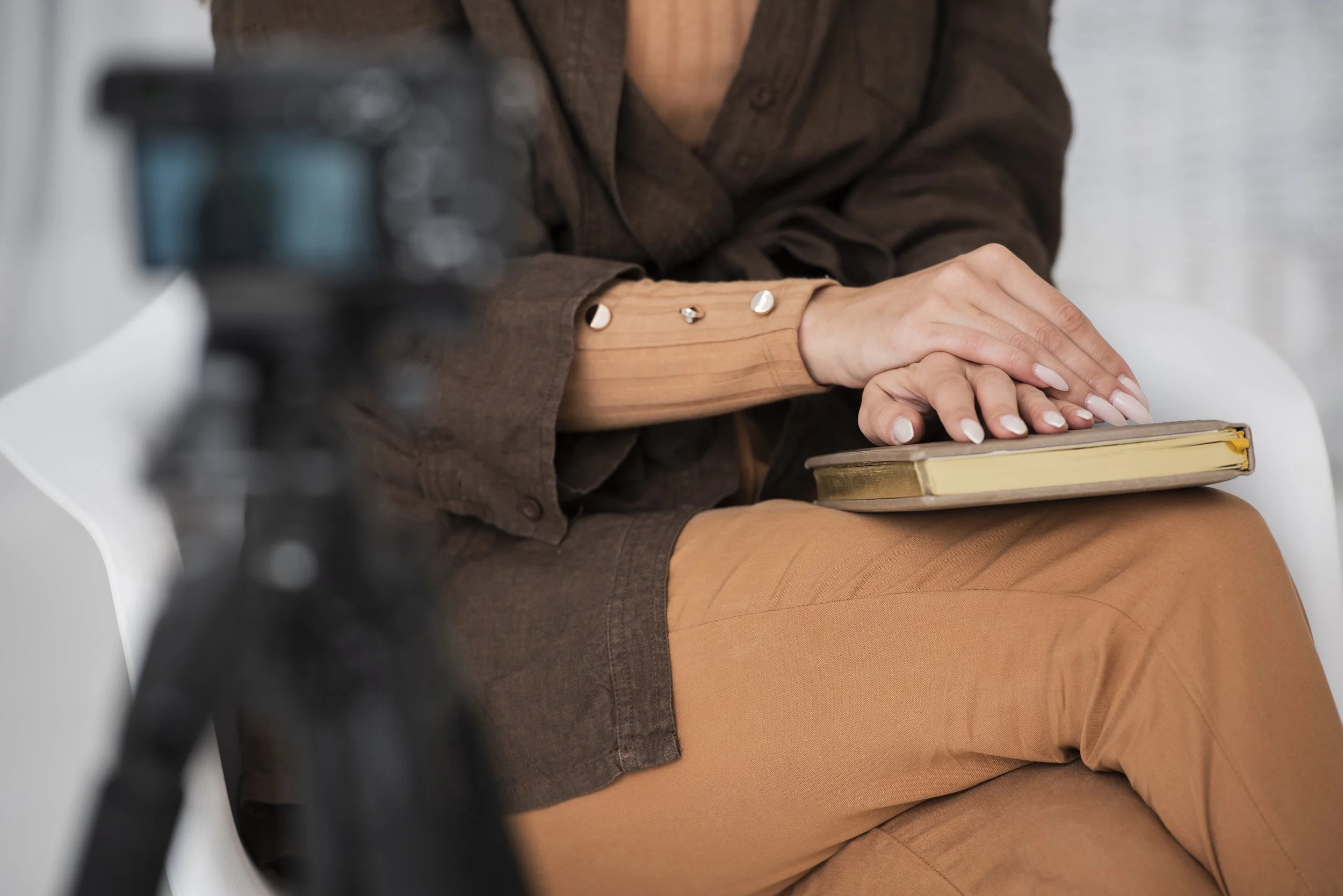 A person sitting on a white chair with a book on their lap, wearing a brown outfit with decorative buttons on the sleeve, and using a camera to record or stream.