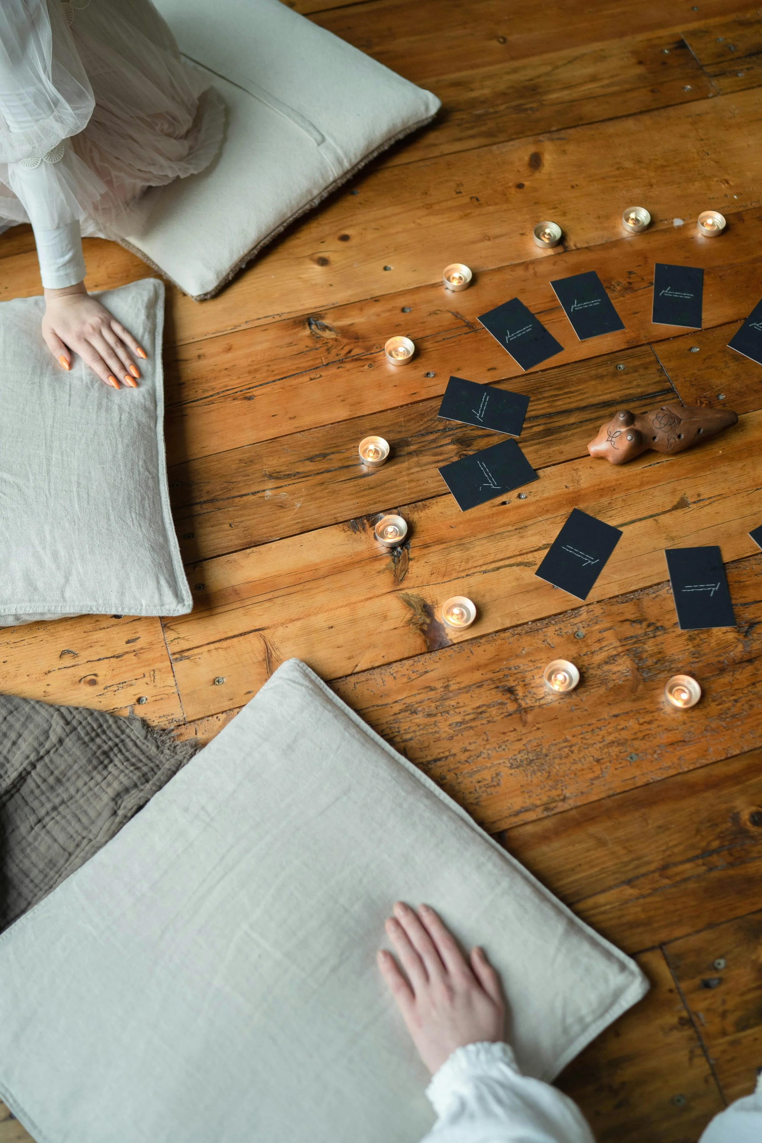 Une personne avec une main posée sur un coussin beige, une table en bois avec des bougies et des cartes, décor minimaliste.