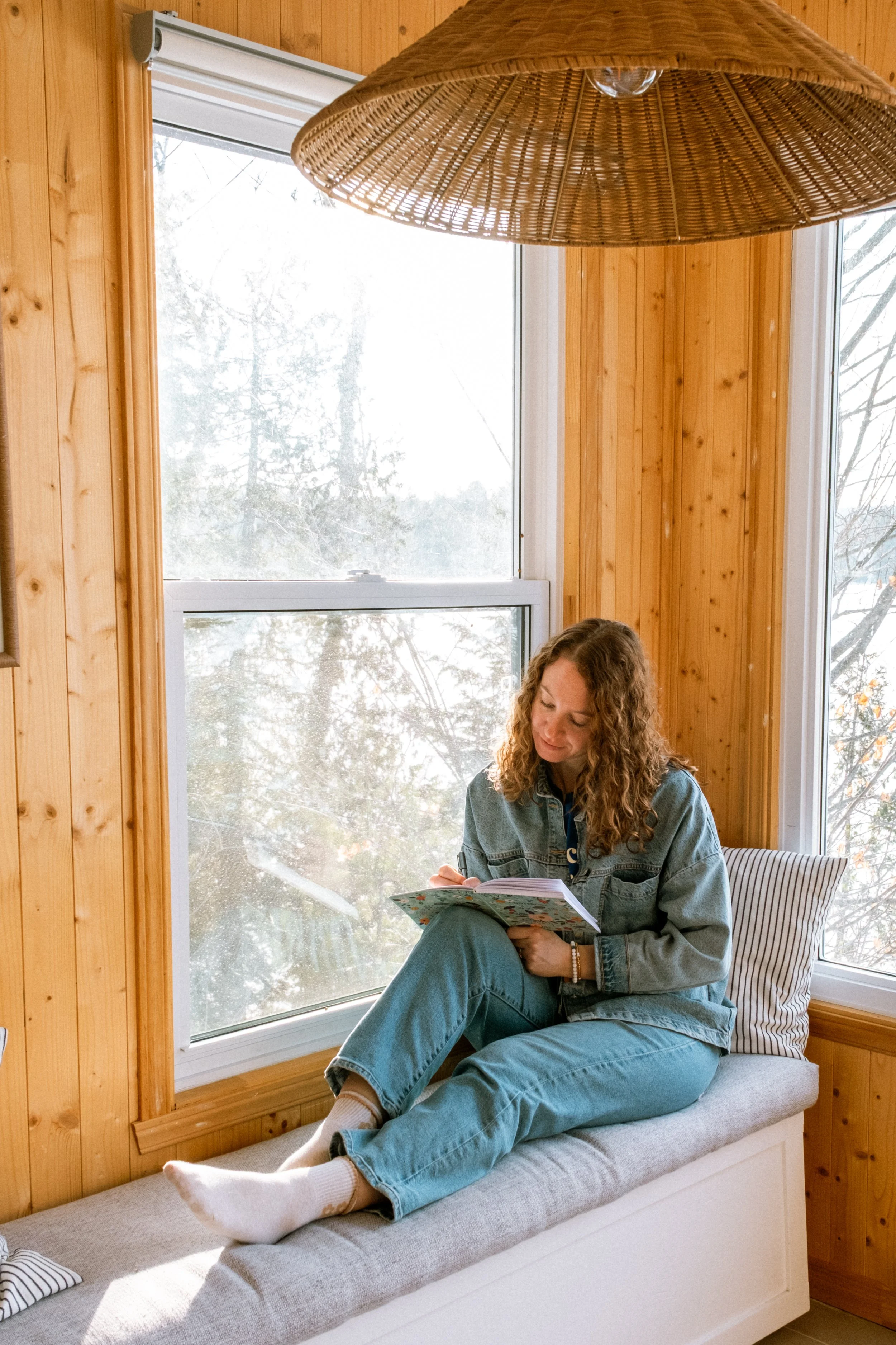 Une femme assise sur un banc de peinture claire près d'une fenêtre avec des murs en bois, lisant un livre.