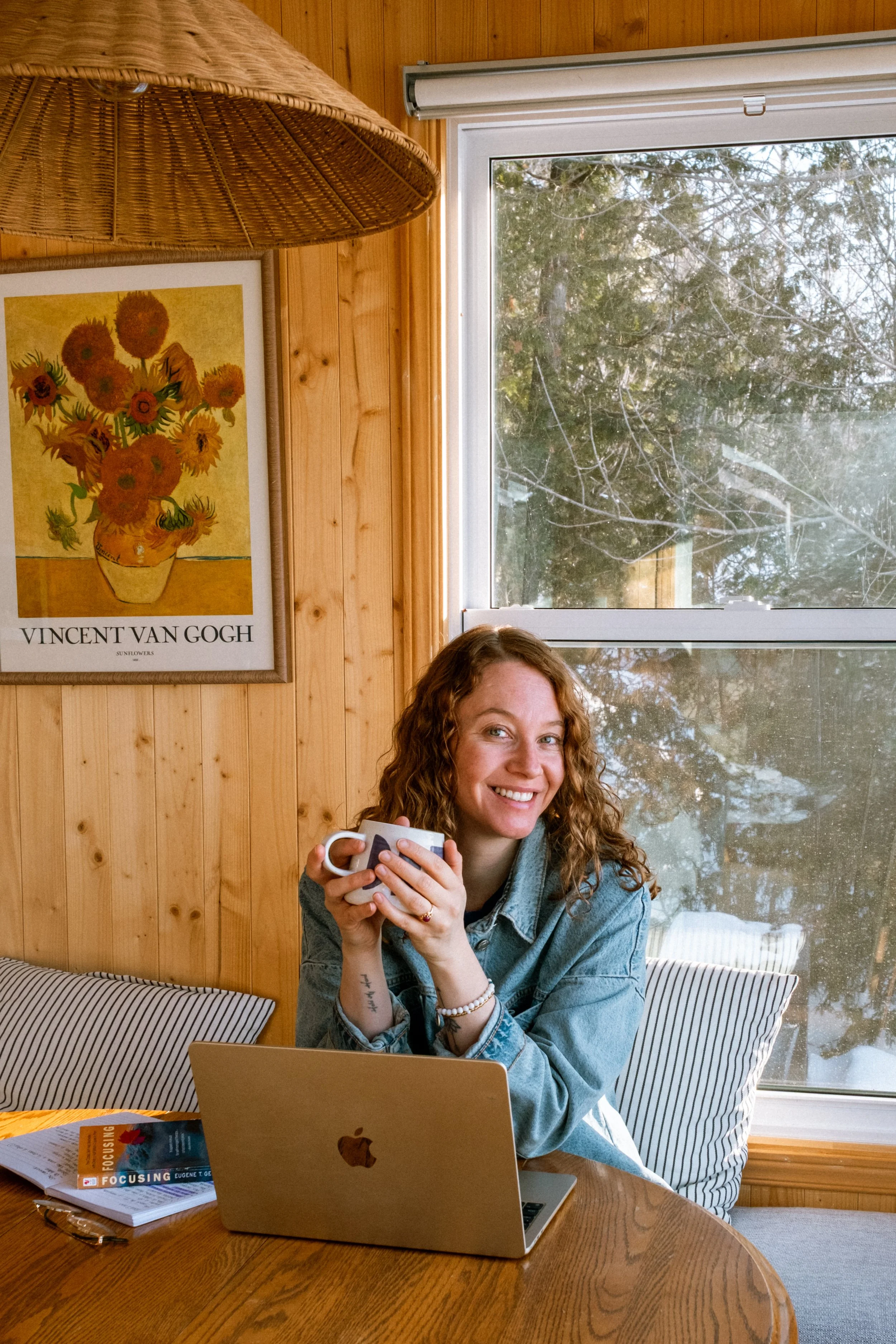 Une femme aux cheveux bouclés assise à une table en bois, tenant une tasse à café, avec un ordinateur portable Apple devant elle, dans une pièce en bois avec un tableau de Vincent Van Gogh et une fenêtre offrant une vue sur des arbres.