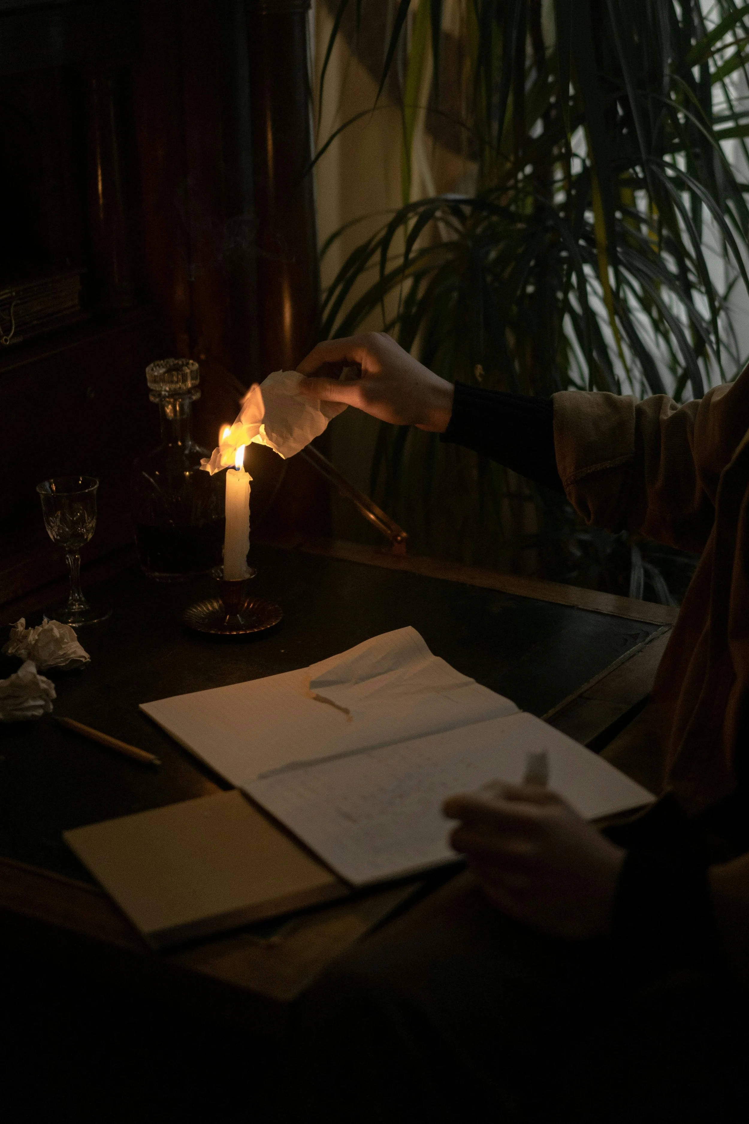 Une personne allume une bougie dans un flacon en verre, à côté d'un verre à vin et de plusieurs pages de papier dans un environnement sombre avec des plantes d'intérieur en arrière-plan.