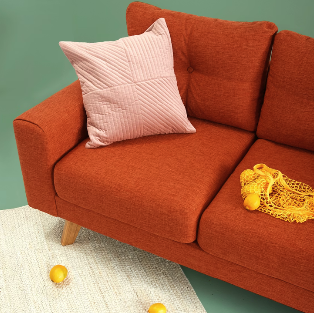 An orange sofa with a pink quilted pillow on it, a yellow plastic bag, two yellow balls on the carpet, and a green wall in the background.