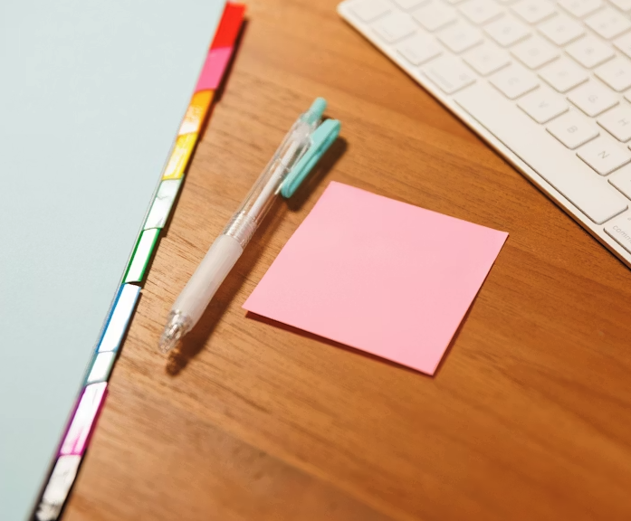 A wooden desk with a light blue notebook, a clear pen with a teal clip, a pink sticky note, and part of a white computer keyboard.