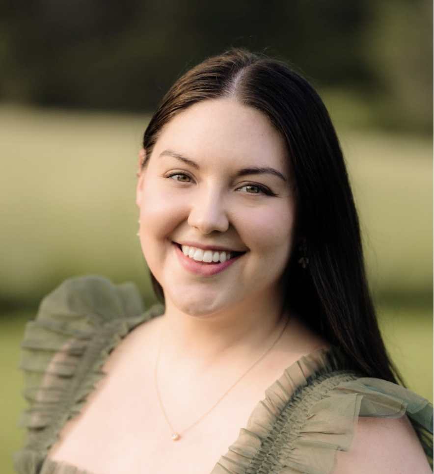 A young woman with long dark hair smiling outdoors in a green field, wearing a ruffled olive-green top and a delicate necklace.
