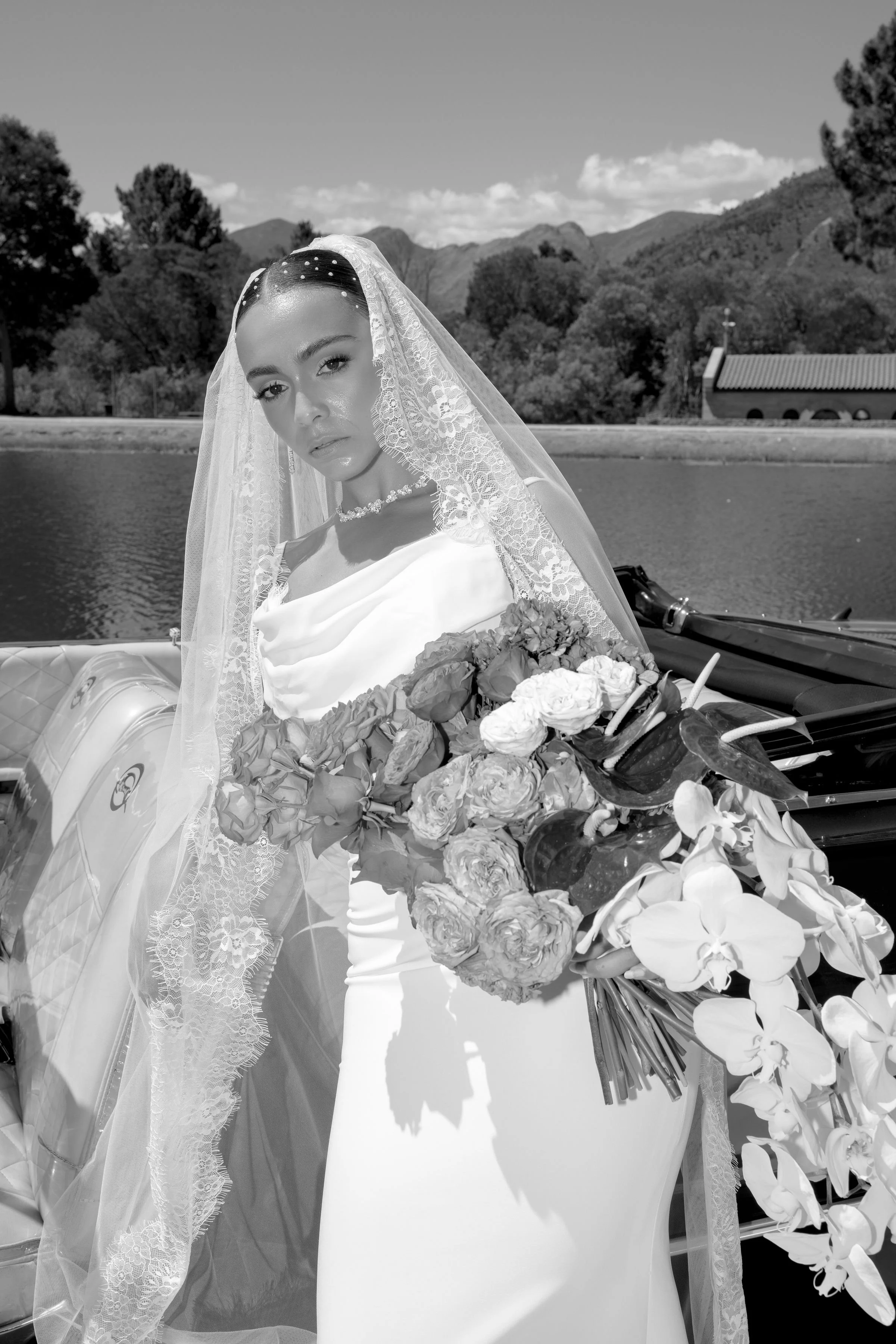 A woman in wedding attire standing in front of a vintage car with a mountain landscape in the background.