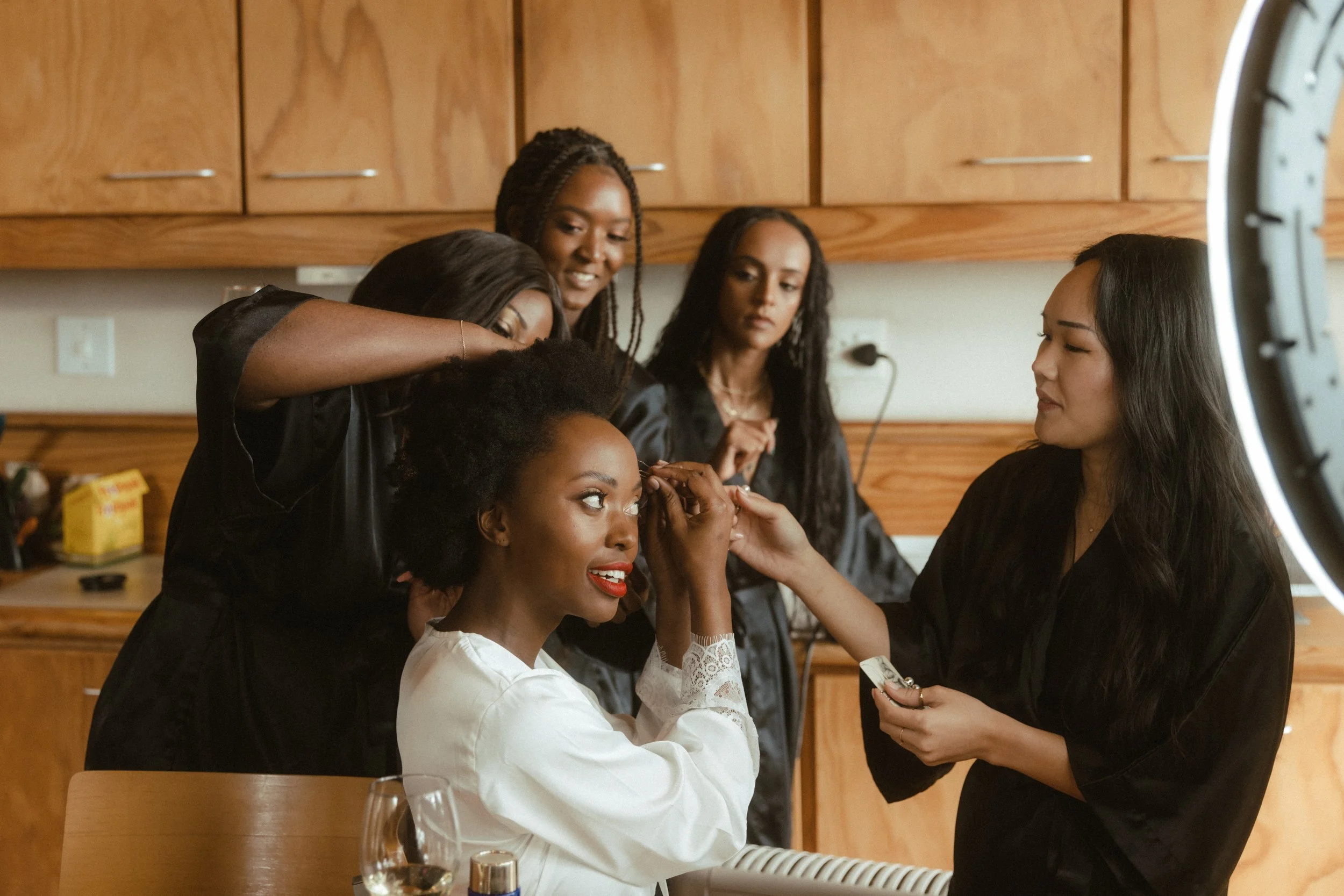 Women getting ready for a photo shoot, with one woman seated and others assisting with makeup and hair, in a room with wooden cabinets.