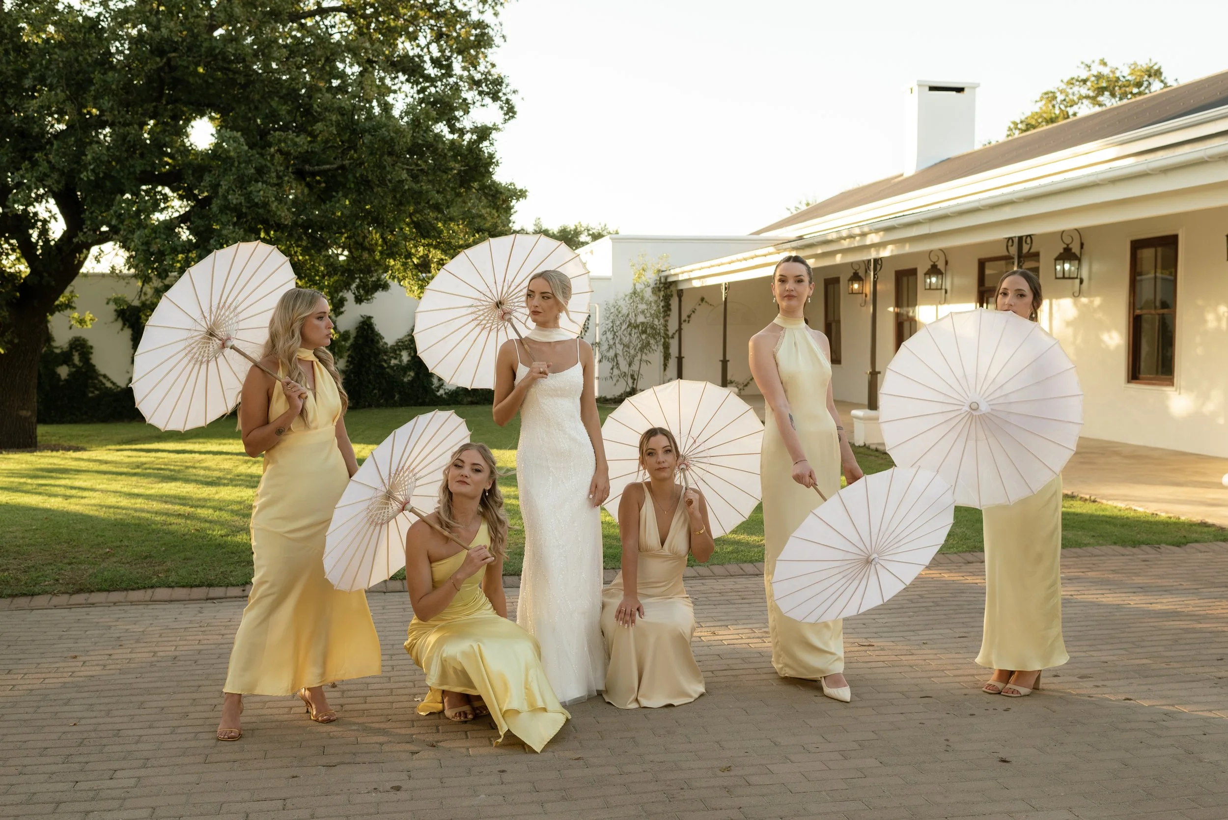 Group of seven women, some standing and some kneeling, wearing yellow dresses and holding white umbrellas, standing outdoors on a brick patio with a white building, trees, and green yard in the background during daytime.