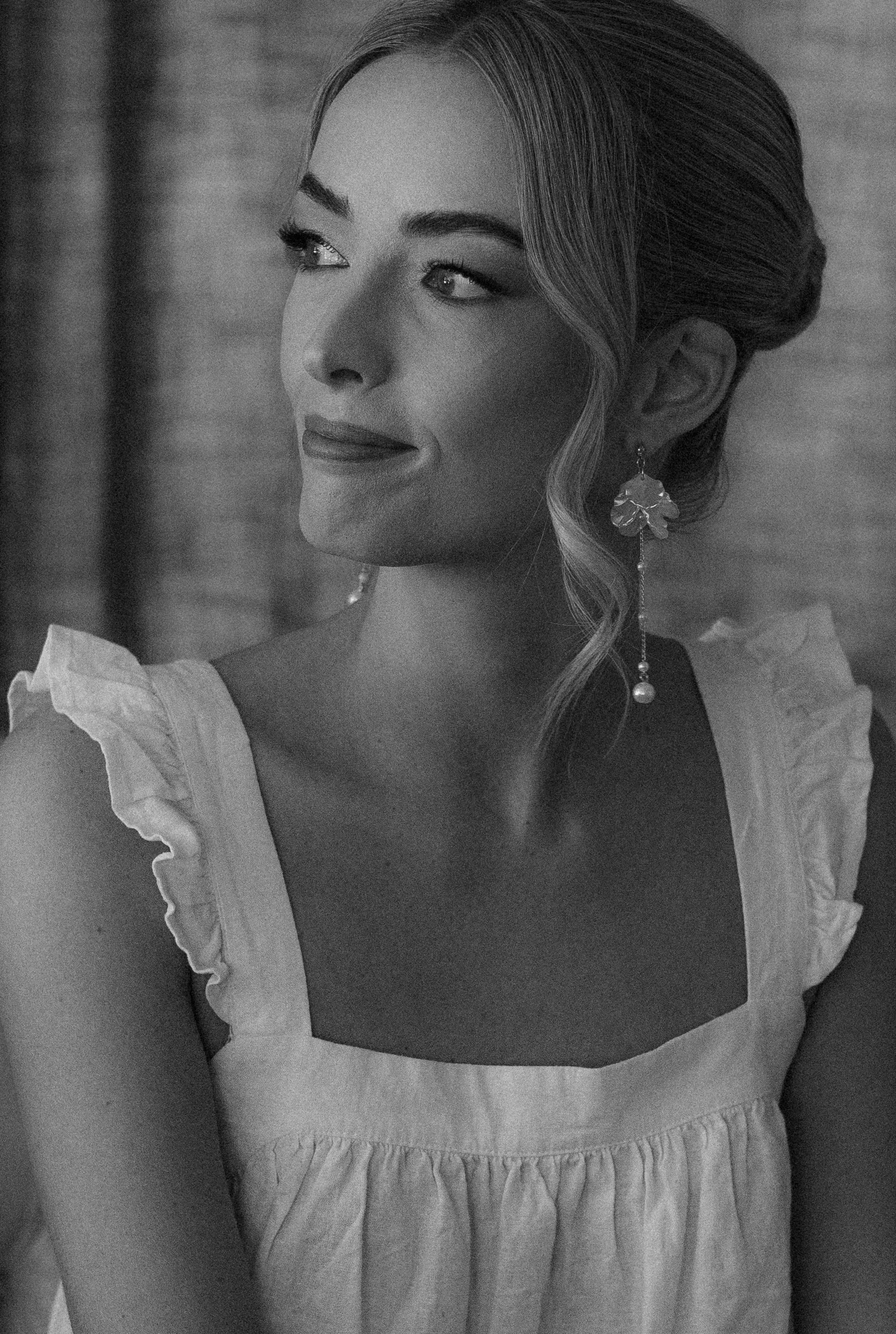 Black-and-white photo of a woman with styled hair, wearing a white ruffled dress and large earrings, looking to her left.